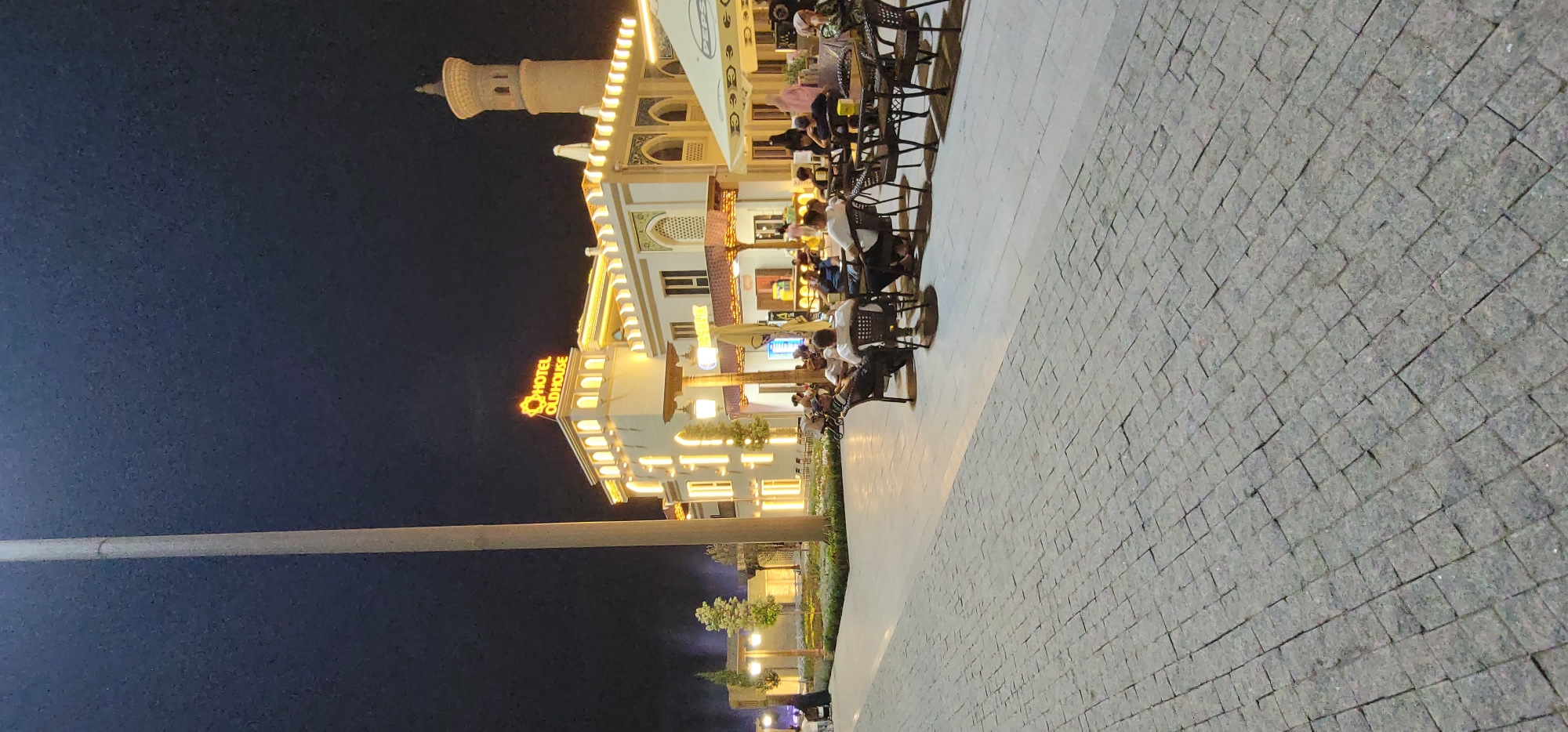 The image shows a nighttime scene outside a building labeled "HOTEL OLDHOUSE." The building is brightly lit with yellow lights, and features a prominent minaret-like structure on the left. In front of the hotel, a paved outdoor seating area is occupied by several people sitting at tables and chairs, engaged in conversation or dining. The ground is paved with stone tiles. The sky is dark, indicating it is nighttime. The overall atmosphere appears relaxed and social.