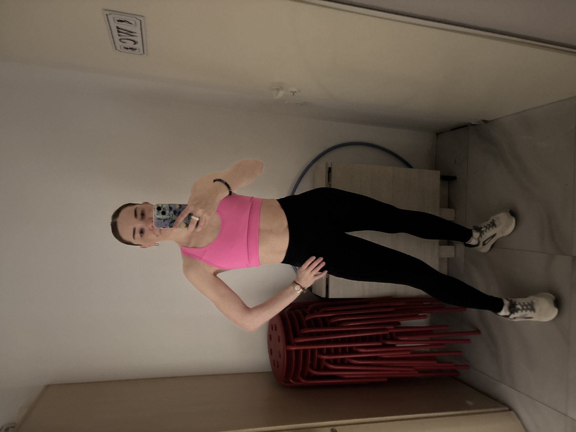 A person is standing in a room, possibly in Unknown, Russia, taking a selfie in a mirror. The person is wearing a pink sports bra and black leggings, with white sneakers. There is a stack of red plastic chairs on the floor near the person, and a blue hula hoop is leaning against the wall behind them. A white door and a sign with black letters are visible on the wall. The lighting appears to be bright and artificial.