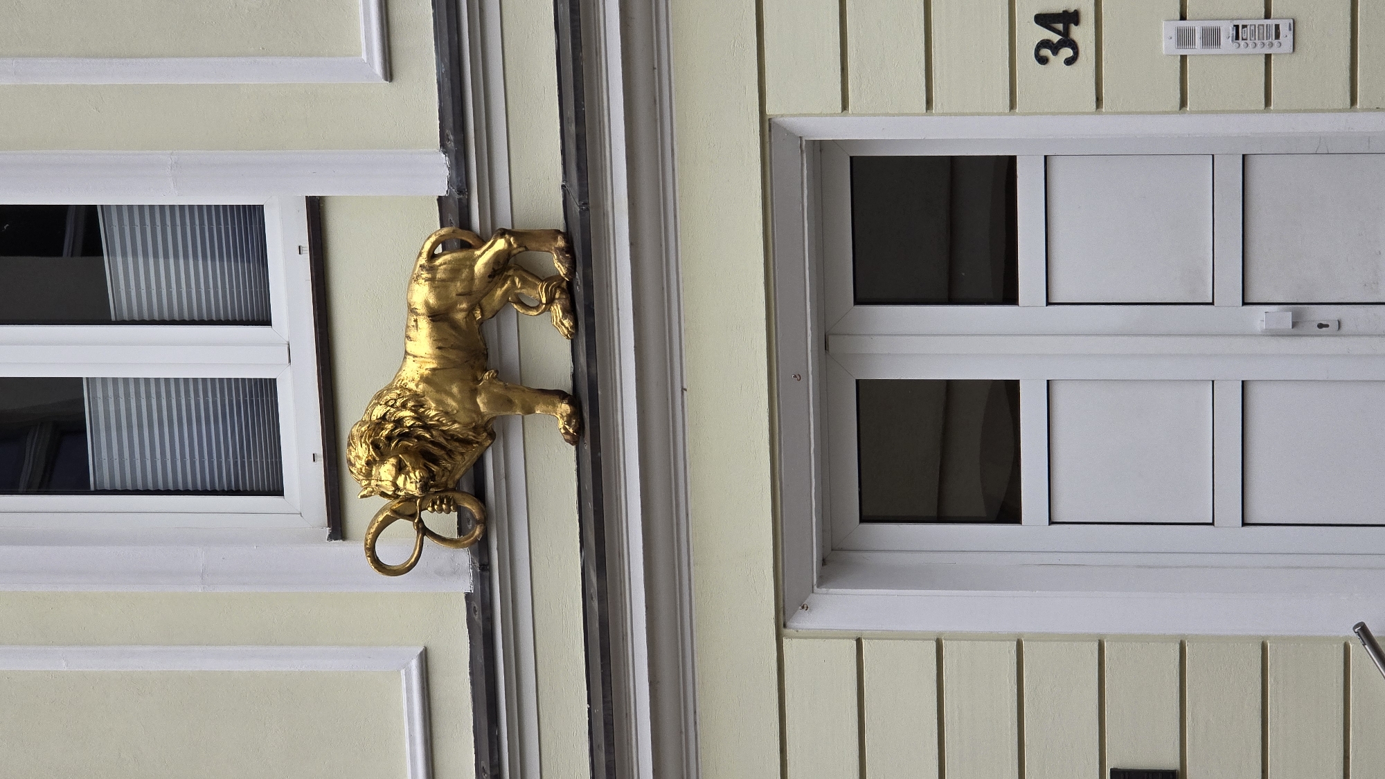 A golden lion sculpture is mounted on the facade of a building in Schwerin, Germany. The lion is depicted in profile, facing left, with its mouth open and holding a coiled rope or chain in its jaws. The sculpture is positioned above a white-framed window with horizontal blinds. Below this window, there is a white double door with glass panels. To the right of the door, a house number "34" is visible, along with a white intercom panel. The building's exterior is painted a pale yellow, with white trim around the windows and door. The architectural style suggests it is an older building.
