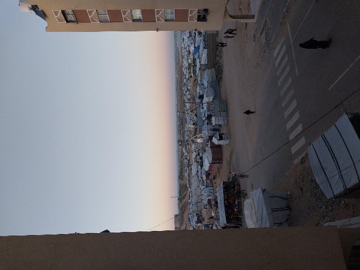The image provides an expansive, high-angle view of a sprawling settlement, likely captured from the upper level of a nearby building during either sunrise or sunset. The perspective looks slightly down and across the scene.
In the immediate foreground, part of a light brown or tan building, presumably the vantage point, is visible on the left side. The top edge of its wall and a few rectangular windows with some sort of protective grates or mesh are seen. One window appears to have a broken pane or a dark mesh behind it. A small satellite dish is mounted on the roof at the far left.
To the right of this building, a wide, unpaved or dirt road runs across the middle of the frame. A clearly painted white pedestrian crossing (zebra crossing) is visible on this road. Several dark, silhouetted figures are present: one person is walking distinctly towards the right side of the frame near the crossing, and a smaller group of two or three individuals are further down the road near the building on the left. A large, dark, indistinct object rests on the far right side of the road, closer to the foreground.
Beyond the road and stretching into the distance on the left, lies a vast and dense settlement. It consists of numerous closely packed, temporary or semi-permanent structures, characterized by their grey and silver tones, suggesting materials like corrugated metal, tarpaulins, or canvas. These structures form a complex, crowded pattern of roofs and walls. Along the roadside, facing the settlement, are smaller, makeshift stalls or shops, one of which appears to display goods. A thin, dark line, possibly a power cable or wire, stretches diagonally from the lower right corner, across the road, and over a section of the settlement.
The settlement extends far into the background, eventually meeting the distant horizon. The sky, which occupies the upper portion of the image, displays a soft gradient. It transitions from a clear, light blue-grey directly overhead to a warmer, delicate peach or pale orange hue closer to the horizon. This sky coloration, combined with the presence of long shadows cast by the figures on the road, indicates the soft, low-angle light of either early morning or late afternoon. The overall impression is that of a quiet, observational moment in a busy, inhabited area.