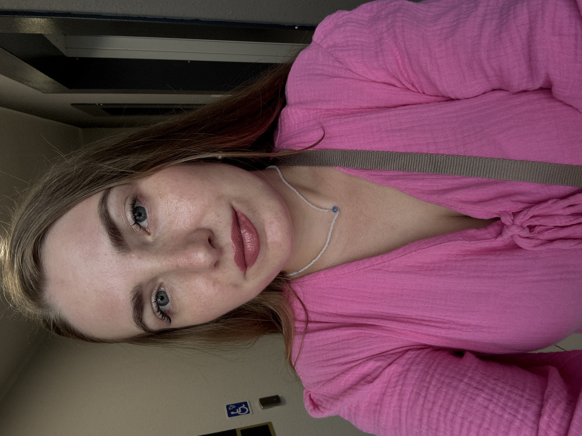 A woman is taking a selfie in what appears to be an indoor hallway. She has light brown hair and blue eyes, and is wearing a bright pink textured top. She is also wearing a simple white beaded necklace with a small blue charm. In the background, there is a wall with a handicap accessibility symbol. The lighting is dim. The woman's face is the primary focus of the image.