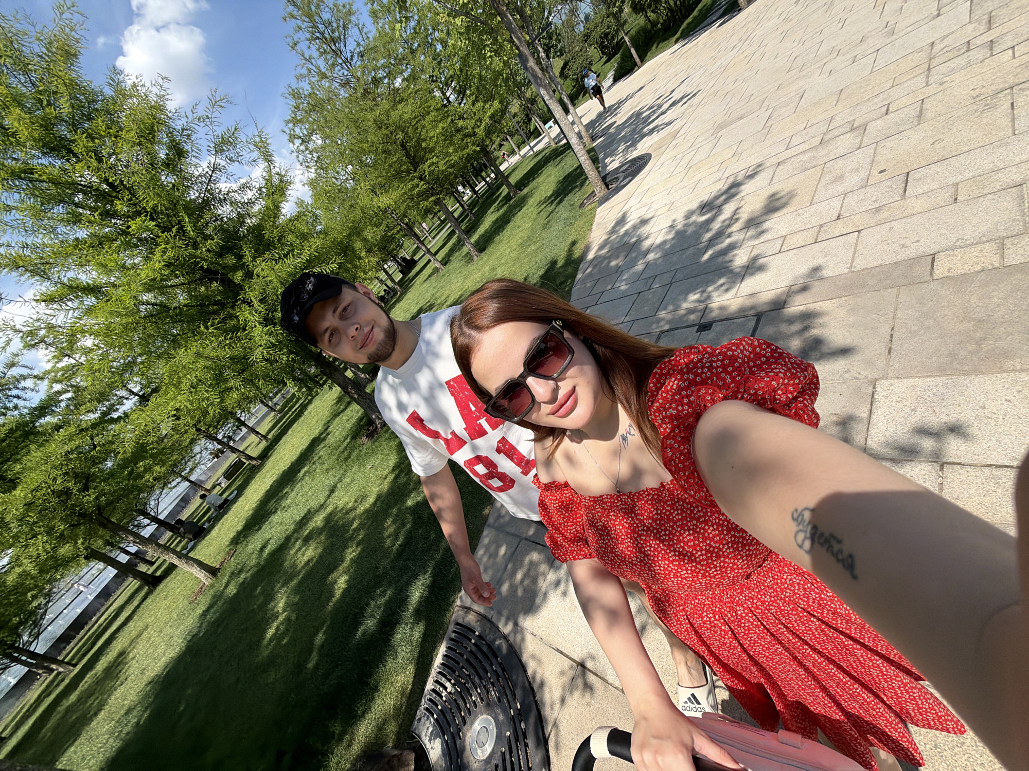 The image depicts a selfie taken by a woman with a man in a modern park on a sunny day. The location is Galitsky Park (Krasnodar Park) in Krasnodar, Russia, identifiable by its distinctive paved pathways, tree species, and a large water feature in the background.
In the foreground, a woman with reddish-brown hair is extending her right arm to capture the photograph. She wears dark sunglasses, a red dress with small white polka dots, and a silver necklace. A tattoo, appearing to read "Dreams" or a similar script, is visible on her right forearm. Her left foot, wearing a white Adidas shoe, is visible next to a white object, and her left hand rests on a black and white striped handle, possibly of a stroller.
Behind her, a man with short dark hair and a light beard is present. He wears a black baseball cap with sunglasses perched on top and a white t-shirt with red lettering, partially visible as "018" or "18." He is looking directly at the camera.
The midground features a wide, light-colored paved pathway composed of rectangular stone blocks. To the left of the woman, a black circular grate is embedded in the ground. Extensive areas of manicured green grass are visible alongside the pathway. Numerous trees with dense green foliage are distributed throughout the park, casting distinct shadows on the ground due to the bright sunlight.
In the background, more trees and grass are visible, leading to a large, calm body of water, possibly a decorative pool or canal. Several indistinct human figures are discernible near the water and on the grass, indicating other park visitors. The sky is clear blue with scattered white cumulus clouds.