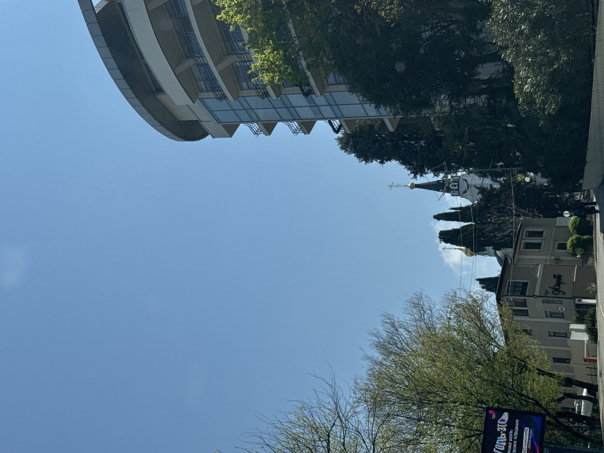 This image captures an urban street scene in Sochi, Russia, under a clear and bright blue sky. The photograph is taken from a low angle, looking upwards towards the buildings and sky.
On the left side of the frame, a modern, multi-story building with a distinctive curved design is prominent. It features numerous balconies and large glass windows, with some green trees partially obscuring its upper sections.
In the mid-ground, towards the center-right, stands an Orthodox Christian church. It has a white facade, a dark steeple, and a golden dome adorned with a golden cross at its peak. The church's traditional architecture contrasts with the modern building nearby.
Various trees with rich green foliage are distributed across the scene, some densely packed, others with sparser, lighter green leaves, suggesting early spring growth. A large tree with visible branches and nascent leaves occupies the bottom-left foreground.
On the far right, another building, possibly commercial, is partially visible. It displays a red circular sign with the number "40" and Cyrillic text on its facade, partially readable as "ФОРУМ" (FORUM). A solitary individual is faintly visible standing on the sidewalk near this building.
At the very bottom left, a section of a large electronic billboard is visible, displaying colorful graphics and Cyrillic text. Visible phrases include "ТОЛЬ-ЭТО" (ONLY-THIS) at the top, followed by "нная боль, твоих близких..." (pain, your loved ones...), and the city name "СОЧИ" (SOCHI) along with "АЛЕКСАНДР" (ALEXANDER).
The scene is bathed in bright daylight, indicated by the strong illumination and the clear, virtually cloudless blue sky that fills a large portion of the frame. The weather is sunny, contributing to a calm and peaceful atmosphere that blends urban development with natural elements and traditional architecture.