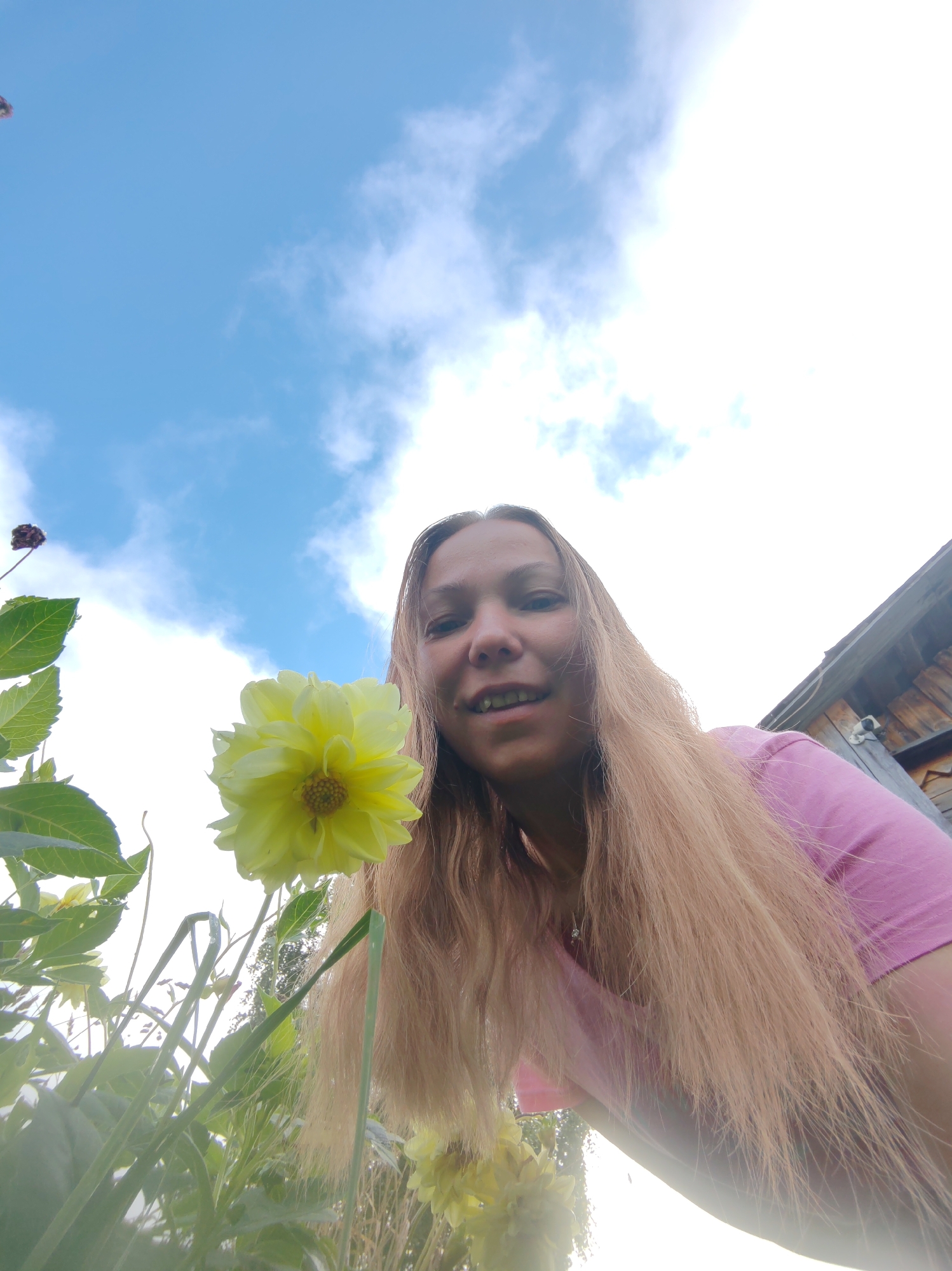 A low-angle, wide shot captures a young woman with long, blonde hair wearing a pink t-shirt, outdoors. She is looking towards the camera with a slight smile, her mouth open. In the foreground, a large, yellow dahlia flower is prominently displayed, with more flowers and green foliage visible. Behind the woman, a weathered wooden structure, possibly a shed or outbuilding, is partially visible. The sky is a bright blue with scattered white clouds, suggesting it's daytime. The overall scene appears to be a garden or rural setting, the location is identified as Chukhlem, Russia.