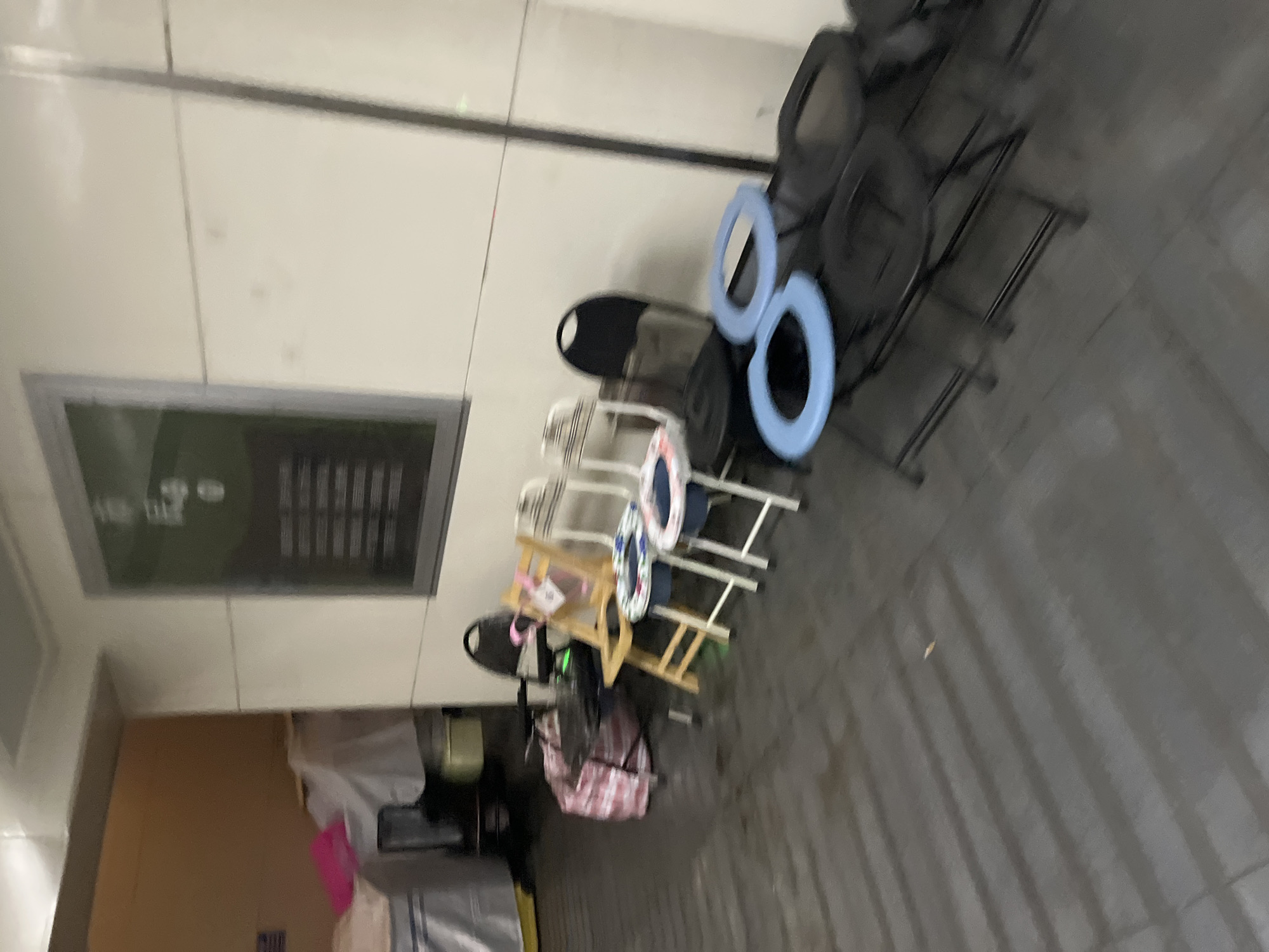 A dimly lit, indoor scene shows a row of chairs and other objects in what appears to be a storage area or hallway in Jiulongpo District, China. In the foreground, several black folding chairs are stacked and arranged, some with blue ring-shaped objects on top, possibly toilet seats. To the left, a light wooden folding chair with a checkered cushion is positioned, next to a large, red and white striped bag. Behind these, a black chair with a curved back is visible. Further back, there are more chairs and what looks like a display case or billboard on the wall. The environment is somewhat cluttered, with some covered items and boxes in the background, and the floor is dark and appears wet in some areas. The lighting is minimal, creating shadows and a sense of disarray.
