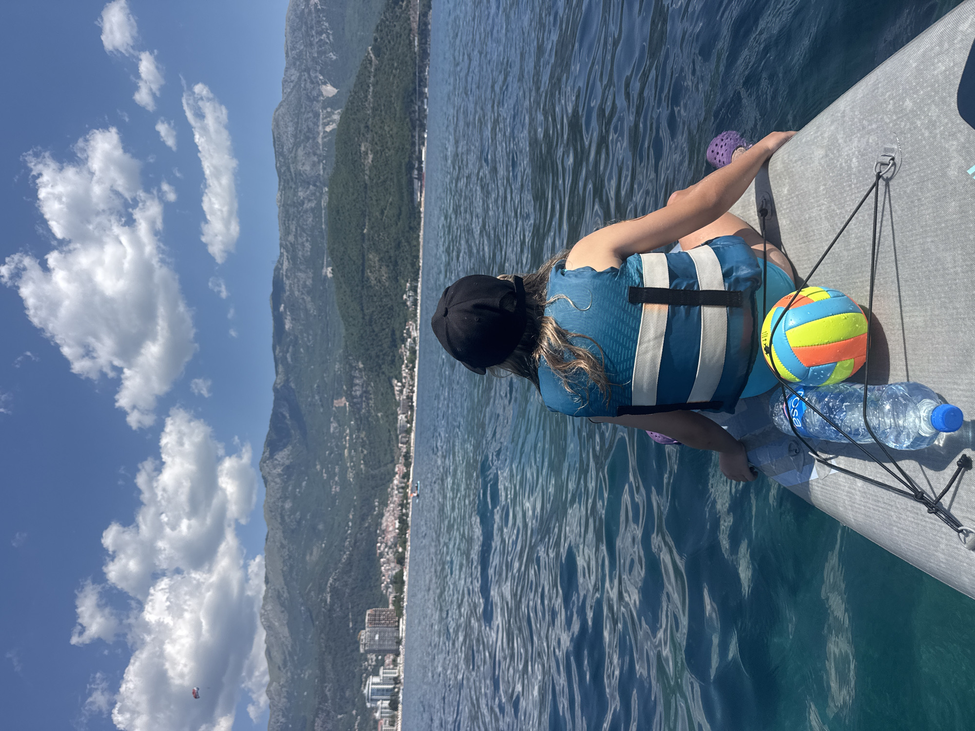 The image shows a young person from behind, sitting on a paddleboard on a body of water. The person is wearing a black cap, a blue life vest with white stripes, and teal swimming bottoms. A colorful ball and a bottle of water are on the paddleboard next to the person. The water is choppy and a deep blue. In the background, there is a coastline with buildings and mountains. Specifically, the skyline and mountainous backdrop are characteristic of Budva, Montenegro. The person appears to be enjoying a recreational activity on the water.