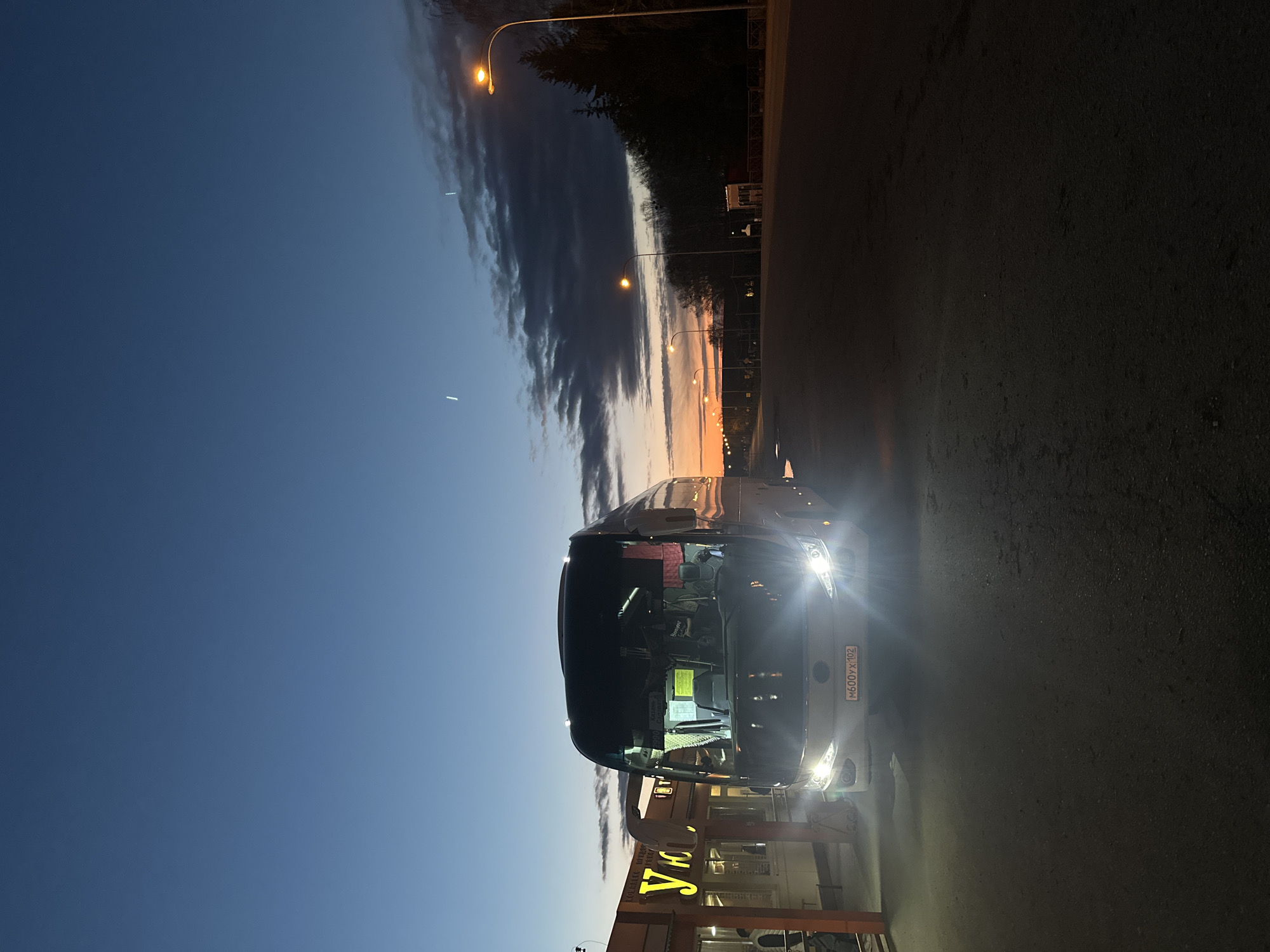 The image, taken during twilight, captures a parked bus at what appears to be a roadside stop in a rural or semi-urban setting, consistent with the Ленино, Russia context. The sky transitions from a deep blue overhead to warm orange and yellow hues at the horizon, with dramatic, dark clouds stretching across. Several streetlights are illuminated along a receding road, casting a warm glow.
In the foreground, a large passenger bus with its bright headlights on is prominent. The bus features a Russian license plate "M600YX 102". Part of a building is visible to the right of the bus, displaying a prominent sign with the Russian word "Уют" (Uют), which translates to "Cozy" or "Comfort." Below this, smaller, less legible Russian text suggests services like a "canteen" (столовая) and "bakery" (выпечка). The ground is dark asphalt.
No people are clearly visible. The overall atmosphere is calm and quiet, characteristic of a travel stop during the early morning or late evening hours.