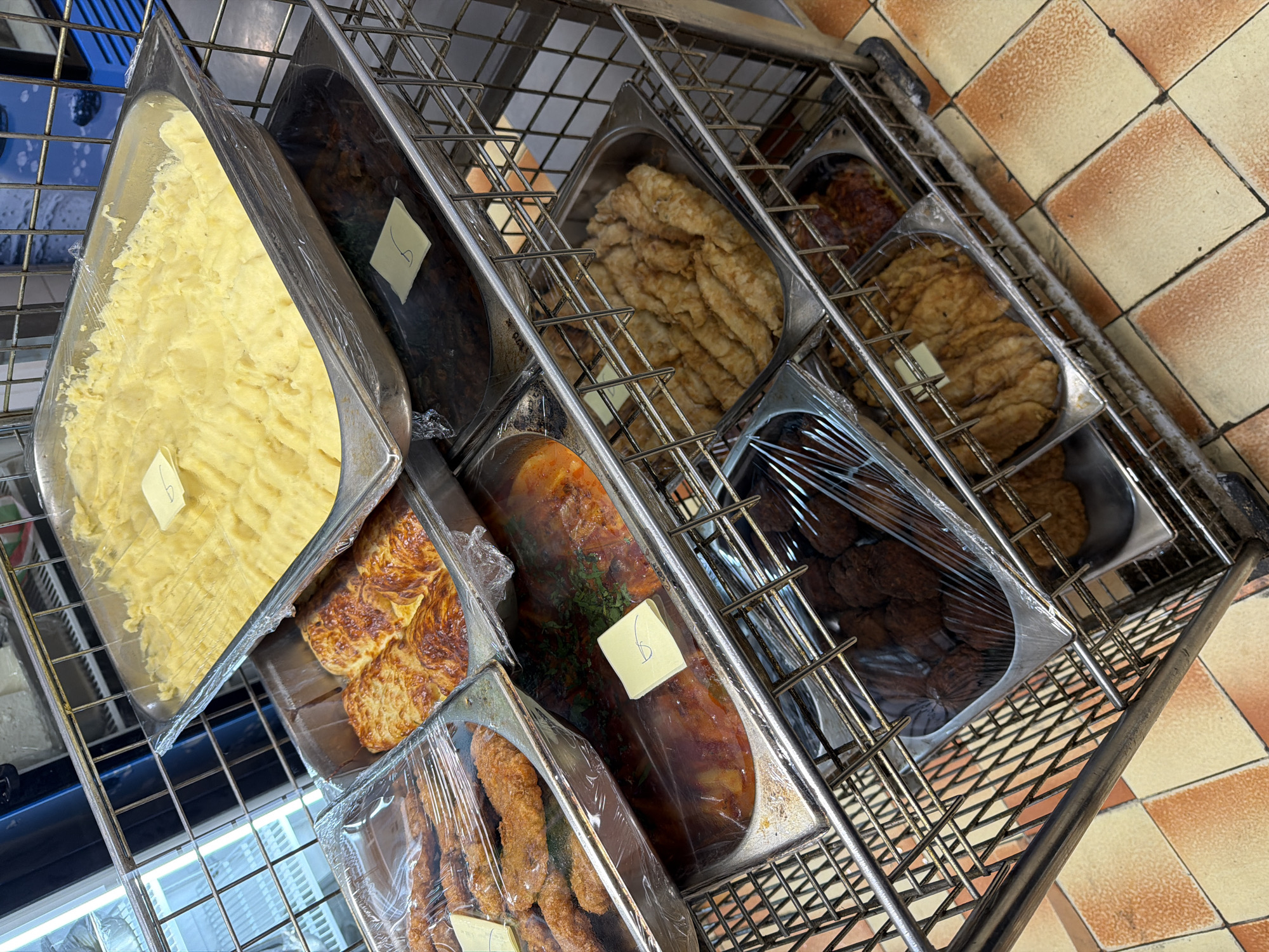 A top-down view shows a metal trolley with various food items in metal trays. The trays are covered in plastic wrap. The food items appear to be mashed potatoes, some kind of dark stew, breaded fish, fried bread or pastries, meatballs, and breaded meat. Small labels are adhered to some of the trays. The trolley rests on a checkered tile floor. The location is Varna, Bulgaria.