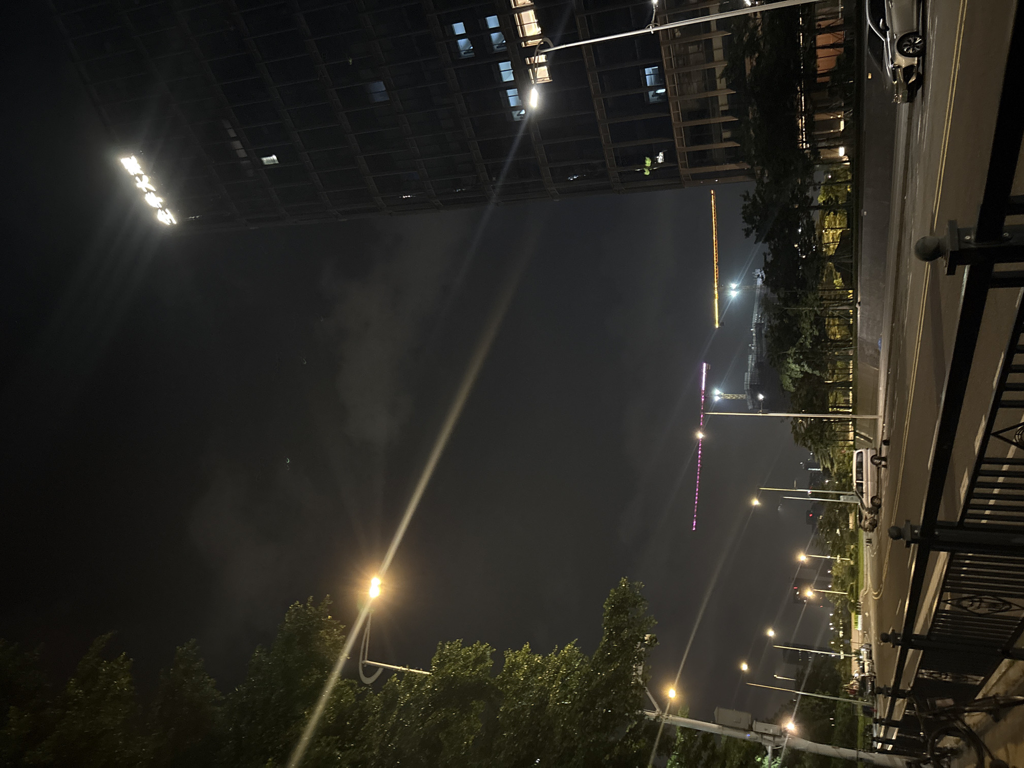 This is a night-time, tilted, eye-level shot of a city scene in Haizhu District, China. The scene is dominated by urban infrastructure and nighttime lighting. On the left, a building with a grid-like pattern of lights can be seen, suggesting an office or residential building. Several bright floodlights are attached to this structure, illuminating the dark sky and casting visible beams. The sky itself appears hazy or foggy, possibly due to pollution or mist, which diffuses the light sources.
In the foreground, trees with dark foliage line a walkway or park area. A street with lane markings and a sidewalk is visible on the right, with several parked vehicles. Streetlights, some with yellow-orange light and others with a cooler tone, are positioned along the road. There is a fence or railing in the lower right corner, indicating a vantage point, possibly a bridge or elevated walkway. Further in the distance, a tall crane with yellow lights is visible, along with more distant buildings and lights that create a typical urban night landscape. A thin crescent moon is faintly visible in the upper right portion of the sky. The overall impression is one of a well-lit, active urban environment at night, with a sense of depth and scale provided by the varying light sources and structures. No people are clearly discernible.