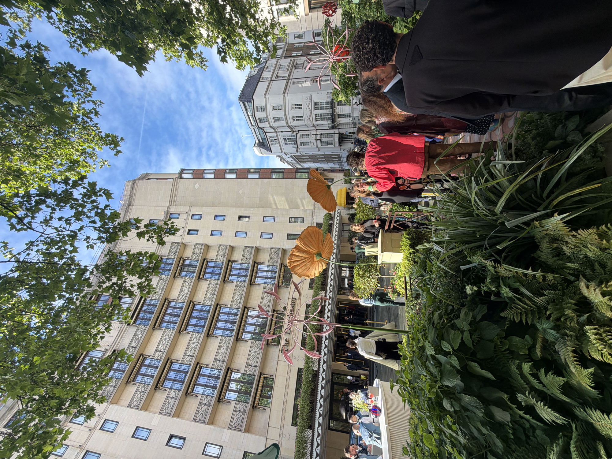 The image captures an outdoor scene in London, United Kingdom, featuring the front of a building, identified by the name "The Beaumont" visible in the picture.
Several individuals are present, some appearing to be in formal attire, such as a black suit jacket. People are standing and sitting at tables outside the entrance of the building. The tables and their environs are decorated with flowers. Overhead, there are several large, decorative flower-like structures that are stylized and brightly colored. The building has multiple windows with decorative metal balconies. The picture includes glimpses of buildings across the street and also shows a blue sky with some clouds visible through some tree branches. The foreground is filled with greenery, including bushes and plants.