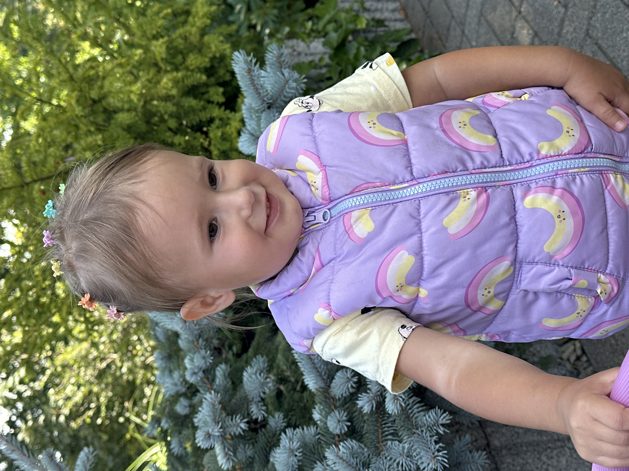 A young child, appearing to be a toddler, is shown from the chest up, outdoors. The child has light brown hair pulled up with small, colorful flower-shaped hair clips. They are wearing a light yellow t-shirt with small cartoon character prints and a lavender puffer vest adorned with a pattern of smiling rainbows. The child is looking to their right with a slight smile and their mouth open. In the background, there are green coniferous trees and out-of-focus green foliage. The ground is paved with gray interlocking pavers. The image appears to have been taken in Wisła, Poland, given the context of other images from the same set, likely in a garden or park setting.