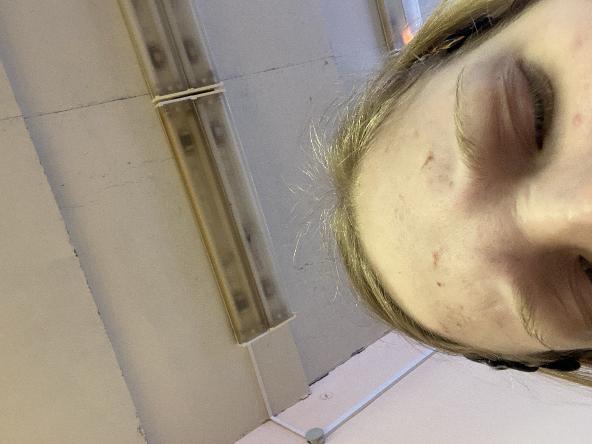 A low-angle photograph shows a person's face in the foreground, angled towards the camera. The person has fair skin, blonde hair, and closed eyes. Behind the person, the background consists of a white ceiling with fluorescent light fixtures. The environment appears to be an indoor space, possibly a commercial or institutional setting. The city and state are unknown.