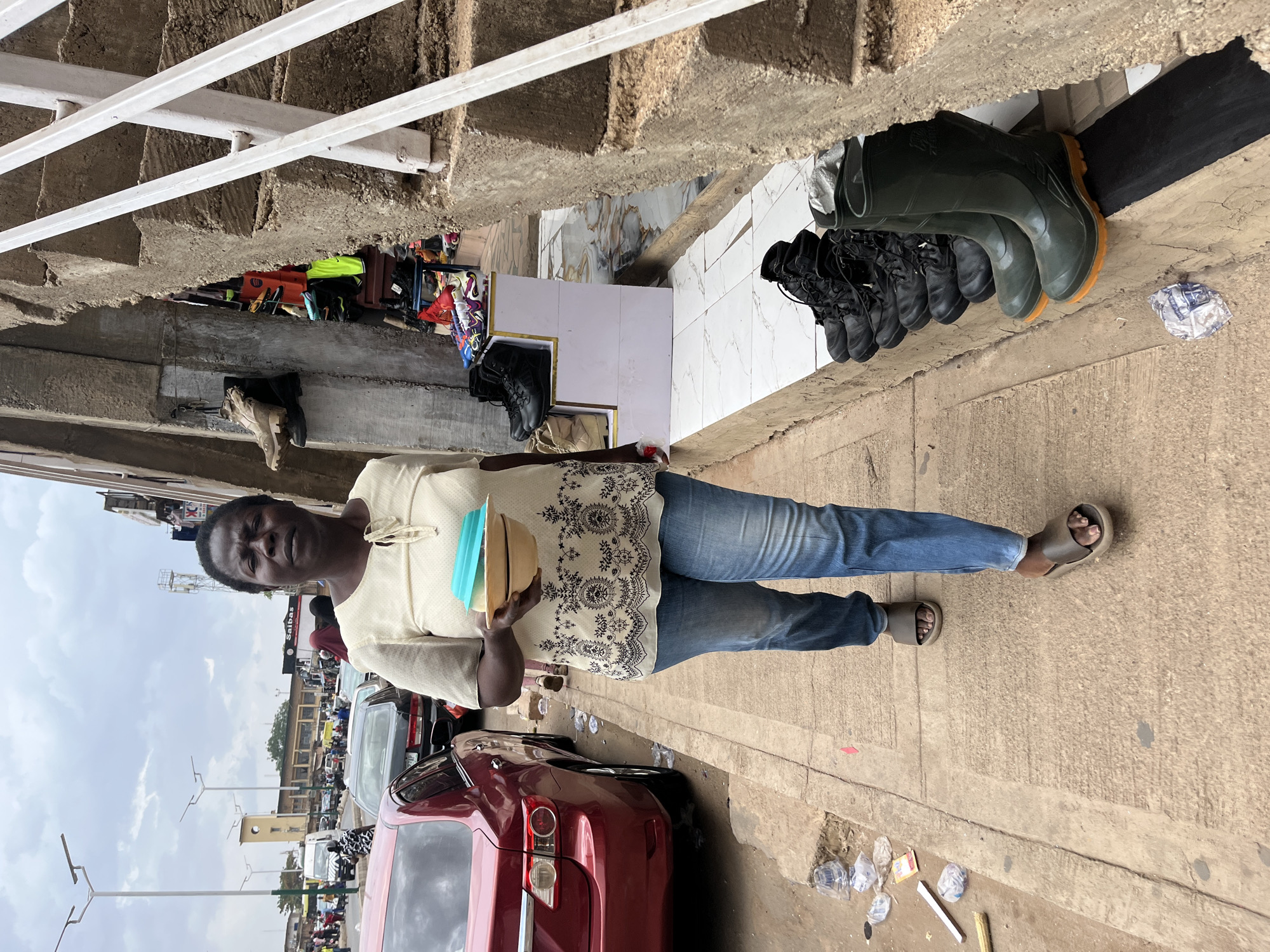 A street scene in Jos, Nigeria. An African woman stands on a sidewalk next to a red car. She is holding blue and tan bowls. Above her are black combat boots and green rubber boots displayed for sale. Debris litters the sidewalk. There are buildings and cars visible in the background. The sky is overcast.