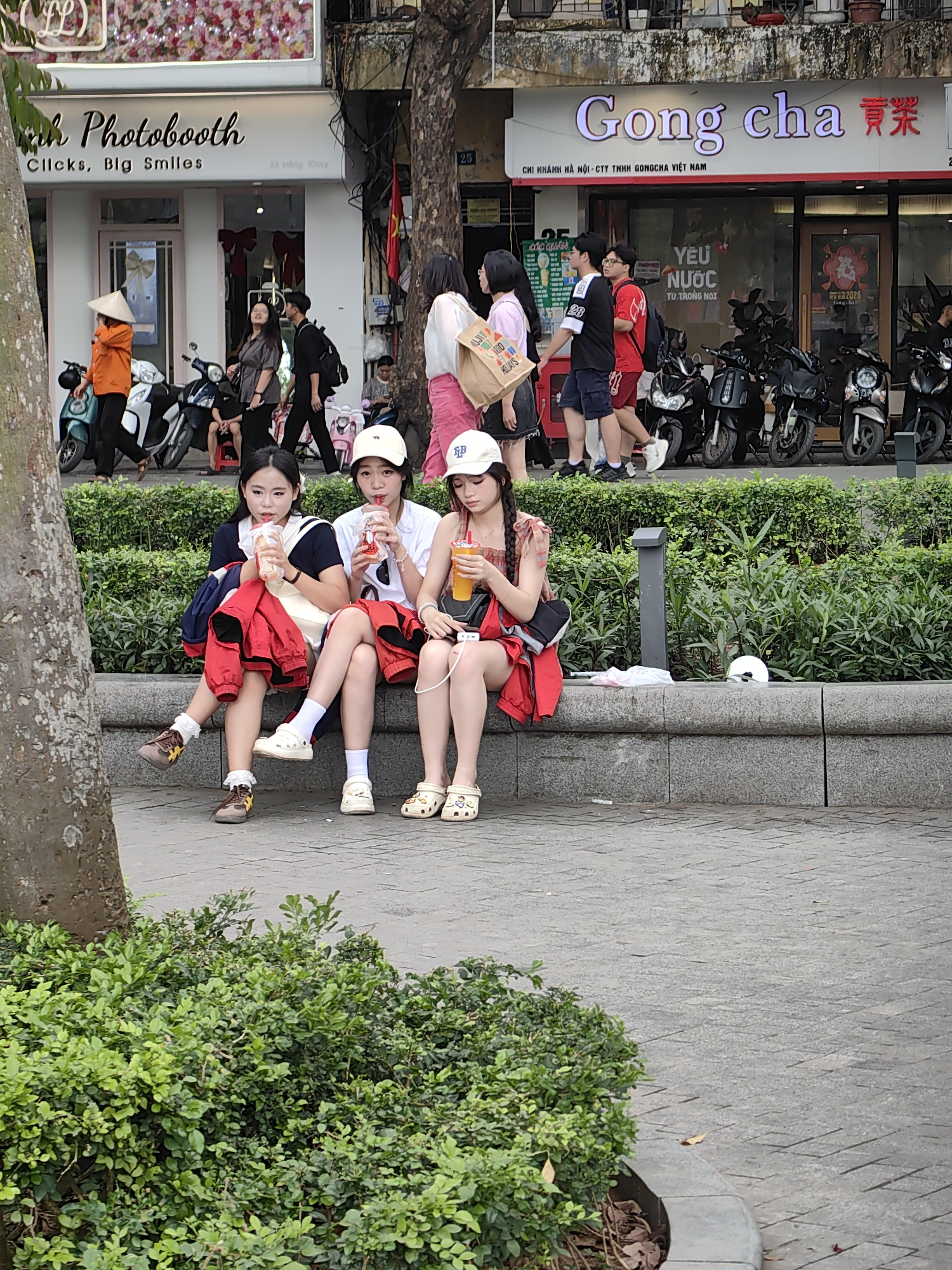 The image depicts a street scene in Hà Nội, Vietnam. Three young women are sitting on a low stone wall, drinking beverages. They are casually dressed, with one wearing a sailor-style top, another a white t-shirt and cap, and the third a patterned top and cap. All three are wearing red jackets or shirts draped over their laps or shoulders. In the background, pedestrians are walking along the sidewalk, and motorbikes are parked in front of two businesses: "Anh Photobooth," and "Gong cha." The Gong cha sign includes the text "CHI NHÁNH HÀ NỘI - CTY TNHH GONG CHA VIỆT NAM" and images of beverages. A tree is on the left side of the frame, partially obscuring the view, and bushes are planted in front of the wall where the women are sitting.
