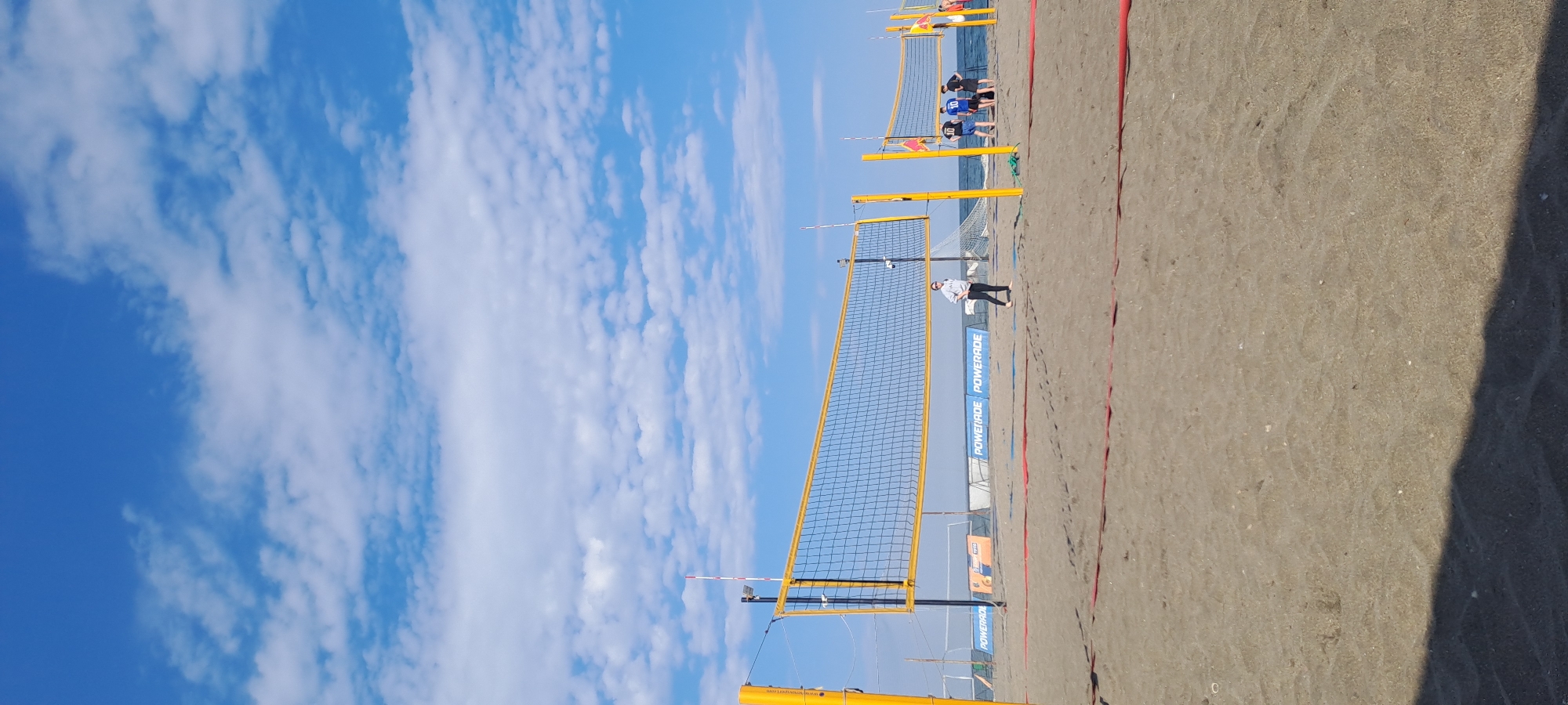 This image depicts a sunny beach volleyball setting in Burgas, Bulgaria, under a bright blue sky dotted with scattered white clouds. The scene is dominated by at least two beach volleyball courts laid out on light brown sand, featuring prominent nets with yellow poles and white boundary tape.
In the foreground, a person wearing a white long-sleeved top and dark pants stands barefoot on the sand, near a net. A red boundary line is visible on the sand court. Further into the background, beyond a second net, a group of several individuals is gathered. Some of these individuals are wearing numbered jerseys, with "10" and "30" being discernible, suggesting they are players or officials. The sea is visible stretching to the horizon behind the courts.
Sponsor banners are affixed to the net poles, clearly displaying "POWERADE" in white text on a blue background. Other faint text on a yellow pole in the foreground includes "www.volleyball.com" and "BOY LEIDERSCHAP BEDRIJF".
The time of day is bright, likely morning or early afternoon, with clear weather conditions. The presence of multiple courts and players suggests a beach volleyball tournament, practice, or recreational event is either ongoing or about to commence. The sandy surface shows various footprints and marks from activity.