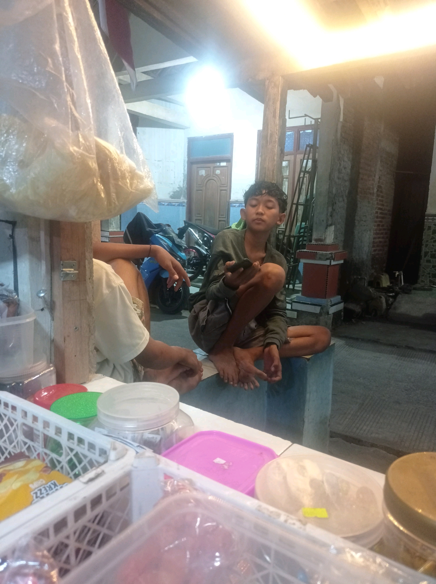 In Babalan, Indonesia, a young male with dark, curly hair sits cross-legged on a concrete ledge, looking down at a mobile phone he holds in his left hand. He is wearing a dark green long-sleeved shirt and dark shorts. His right leg is bent, with his foot resting on the ledge, while his left leg is extended.
To his left, another person, only partially visible, is seated. This individual is wearing a light-colored t-shirt and appears to be holding their left hand in a resting position.
The environment suggests an outdoor setting, possibly a roadside stall or shop. A large, translucent plastic bag filled with a light-colored substance hangs from above in the foreground, partially obscuring the scene. In the background, a blue motorcycle is parked, with a black cover draped over its rear. There are also wooden structures and a brick wall visible.
On the ledge in front of the individuals, various items are arranged on a white surface. These include white plastic baskets containing packaged snacks, colored lids (red and green), and clear plastic containers with lids. The lighting indicates it is either dusk or nighttime, with artificial lights illuminating the area.