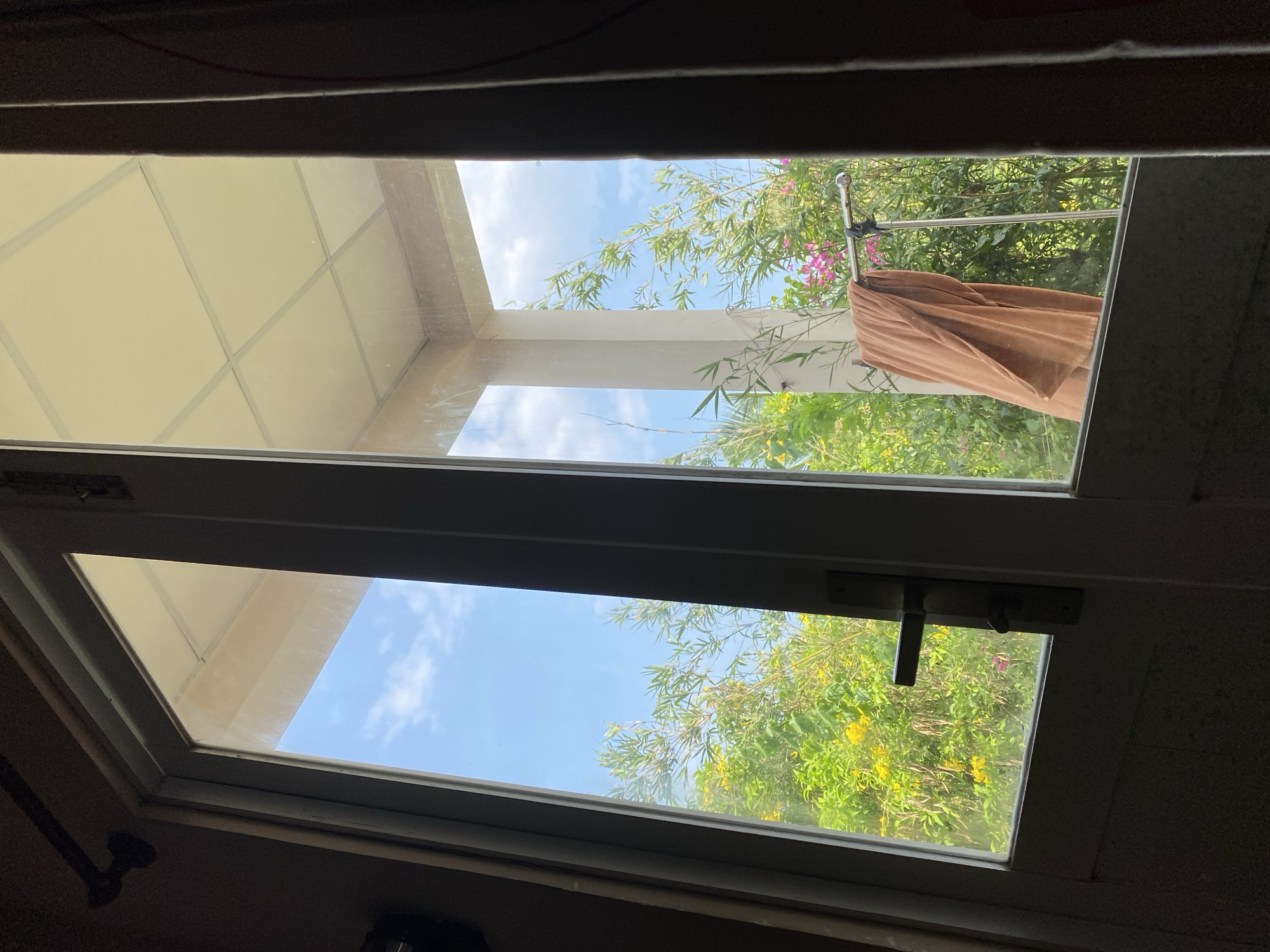 The image shows a view through a glass door into an outdoor area. The door has a dark frame and a silver handle. Outside, there is a patio with a light-colored ceiling composed of square panels supported by pillars. Lush green foliage with yellow and pink flowers is visible. A brown towel is draped over a metal drying rack. The sky is blue with scattered white clouds. The location is identified as Mũi Né, Vietnam.