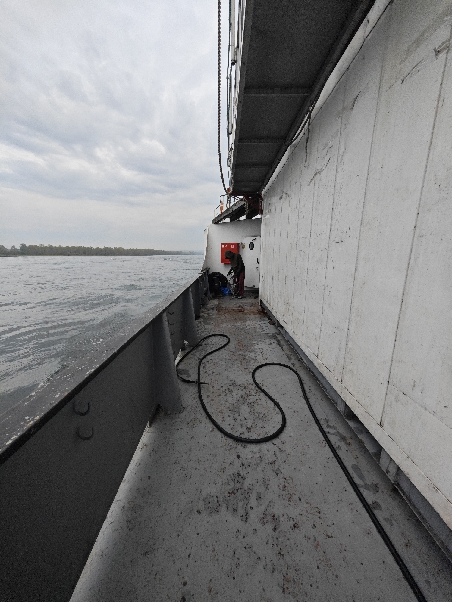 The image depicts the deck of a vessel on a body of water, likely a river, given the tree-lined shore visible in the distance. The sky is overcast with a layer of gray clouds. The deck itself is made of a textured concrete material, with a dark gray metal railing running along the edge. A black hose is coiled and stretched across the deck. To the right, a large, white, rectangular structure, possibly a cargo container or a portion of the ship's superstructure, dominates the scene. The wall of this structure is marked with faint, scratched lines. In the middle ground, a person wearing a hooded jacket and red pants is attending to some equipment on the deck. They appear to be holding a tool and looking down. The water is choppy, indicating movement of the vessel. The overall scene suggests an operational environment on a ship, possibly at anchor or underway. The location is identified as Ермолаево, Russia.