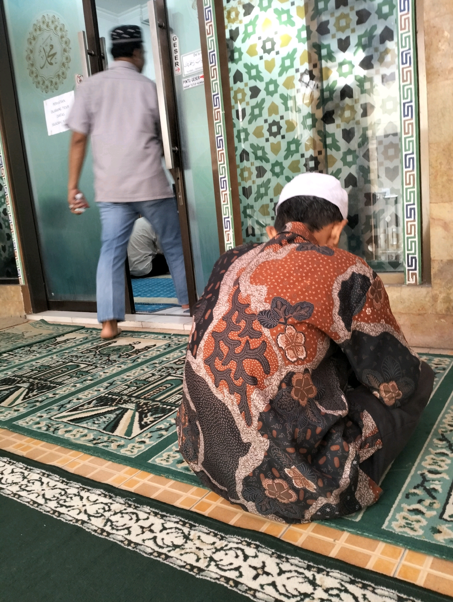An interior shot captures a man in a white prayer cap and a patterned batik shirt kneeling on a mosque carpet. He is facing away from the camera, towards a glass door with decorative green and gold patterns. To his left, another man in a grey shirt and blue jeans walks away from the door, holding a bottle. The floor is covered with a green carpet featuring a geometric pattern. The environment suggests a place of worship, likely a mosque, located in West Jakarta, Indonesia, indicated by the cultural attire and architectural elements.