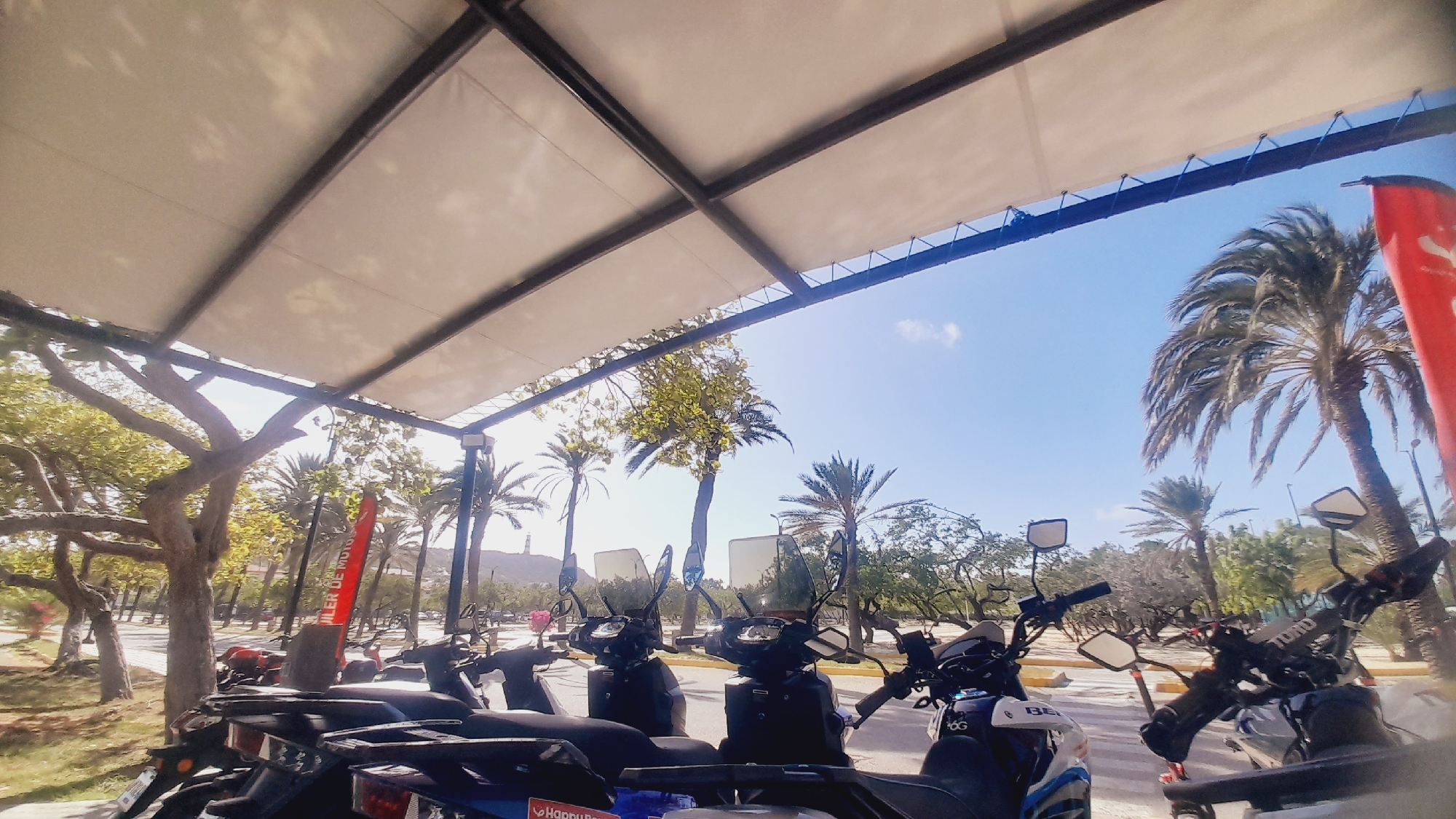 The image shows a view from under a white canopy, looking out at a row of parked scooters or motorcycles. Several palm trees and other lush greenery are visible in the background, along with a bright blue sky. A red flag with white lettering is prominently displayed on the right side of the image. The scooters appear to be lined up in a rental or parking area. The scene is well-lit by sunlight, suggesting it is daytime.