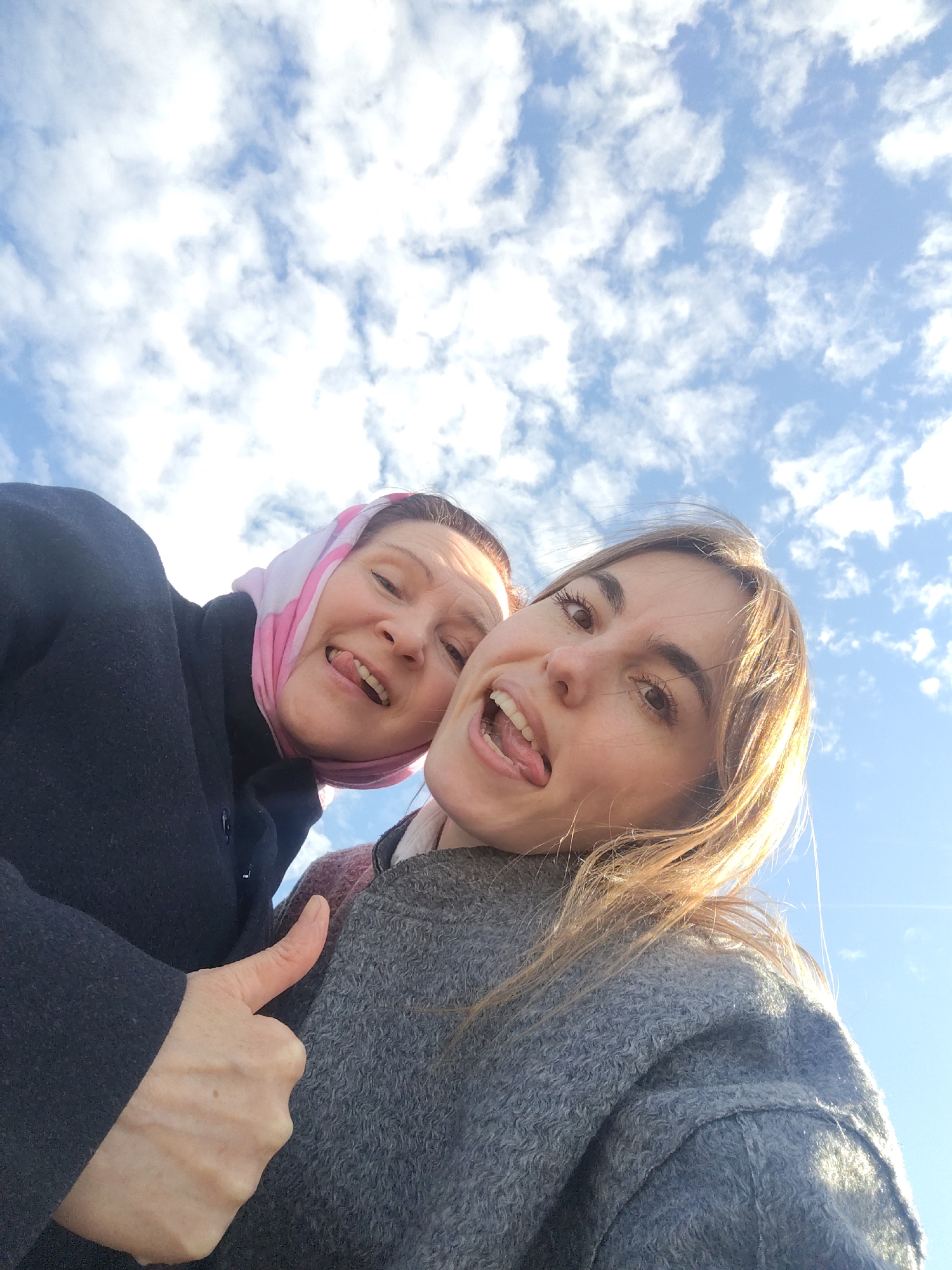 Two women pose for a selfie, sticking out their tongues and looking upwards. The woman on the left wears a dark coat and a pink headscarf. The woman on the right has long brown hair and wears a grey coat. In the background is a blue sky with white clouds. There is no indication of the location in Sarapul, Russia.