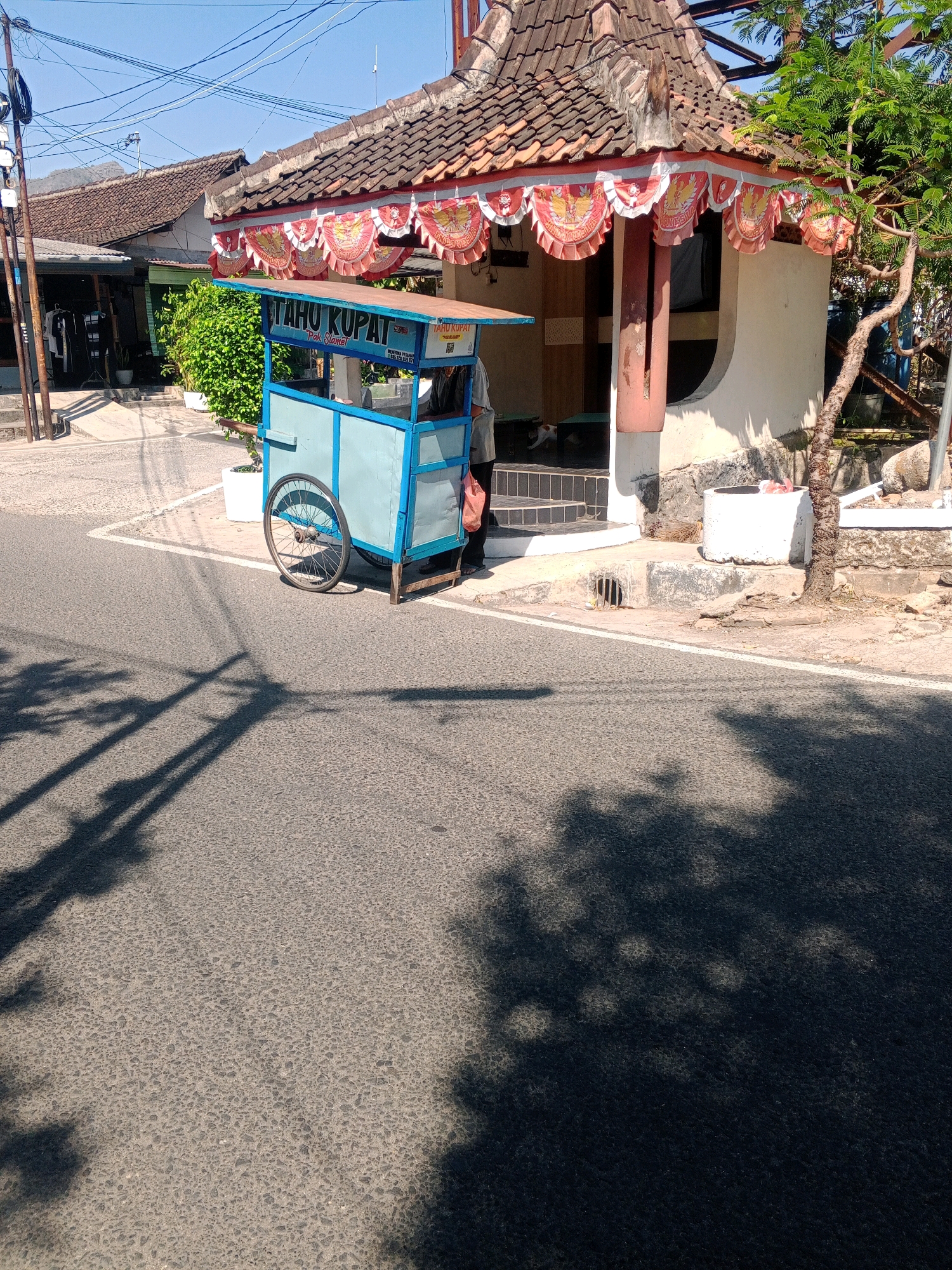 A light blue food cart with a banner reading "TAHU KUPAT PAK SLAMET" is parked on the side of a paved road in Wonogiri, Indonesia, near a small building with a tiled roof and red and white decorative banners. A person in a gray shirt stands behind the cart, holding a pink plastic bag. The scene is illuminated by bright sunlight, casting long shadows across the road and sidewalk. A tree with green leaves is visible to the right of the building.