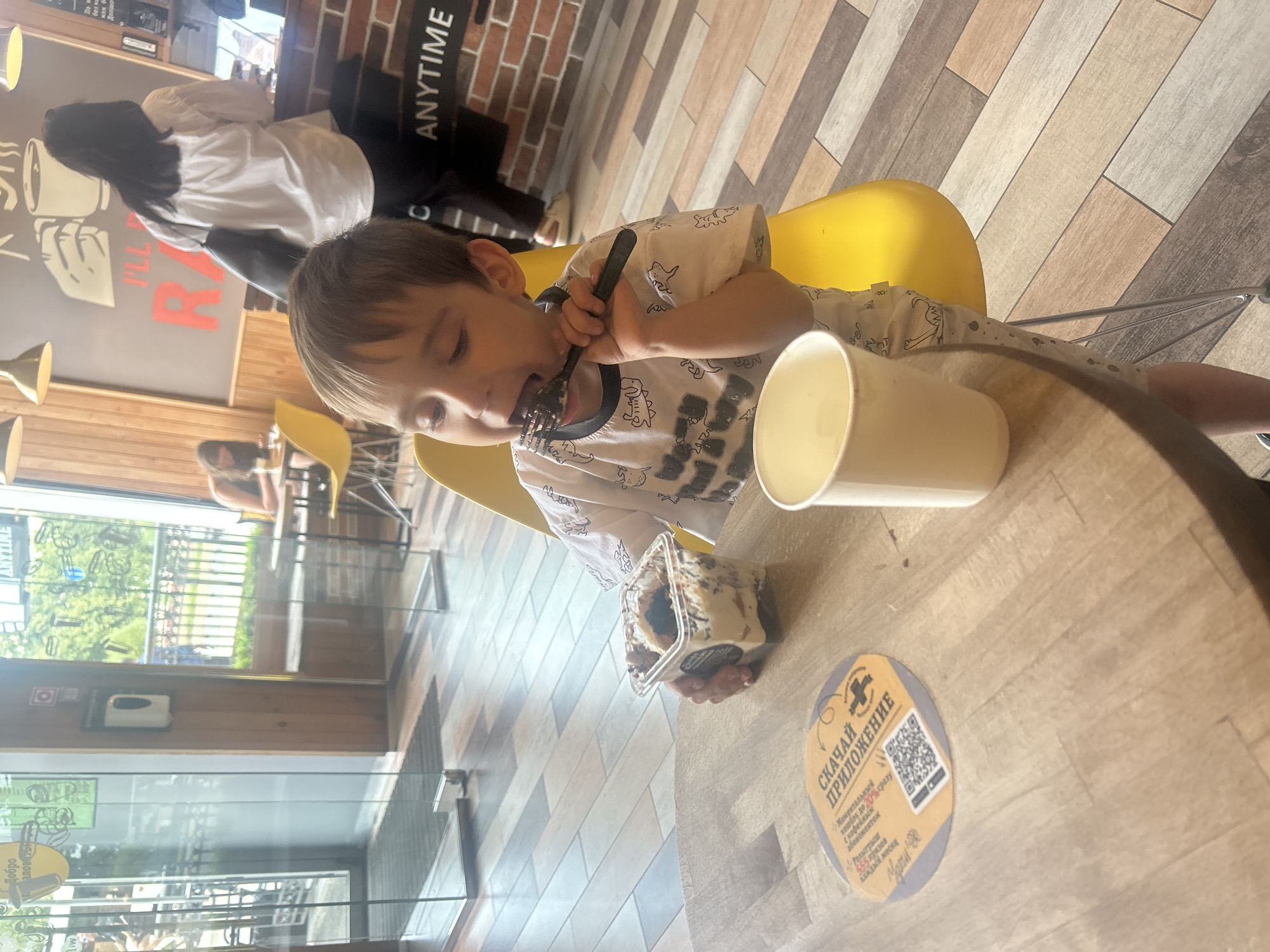 In a casual eatery, a young boy sits at a wooden table, eating dessert from a plastic container with a fork. A white paper cup sits to his right, and a round, orange and yellow coaster with a QR code and Russian text is to his left. The boy wears a white t-shirt with a dinosaur print. In the background, several people are visible. One woman stands at a counter, and another woman sits at a table. The eatery features wooden paneling on the walls and a patterned floor. The overall environment suggests a public place, possibly a cafe or restaurant.