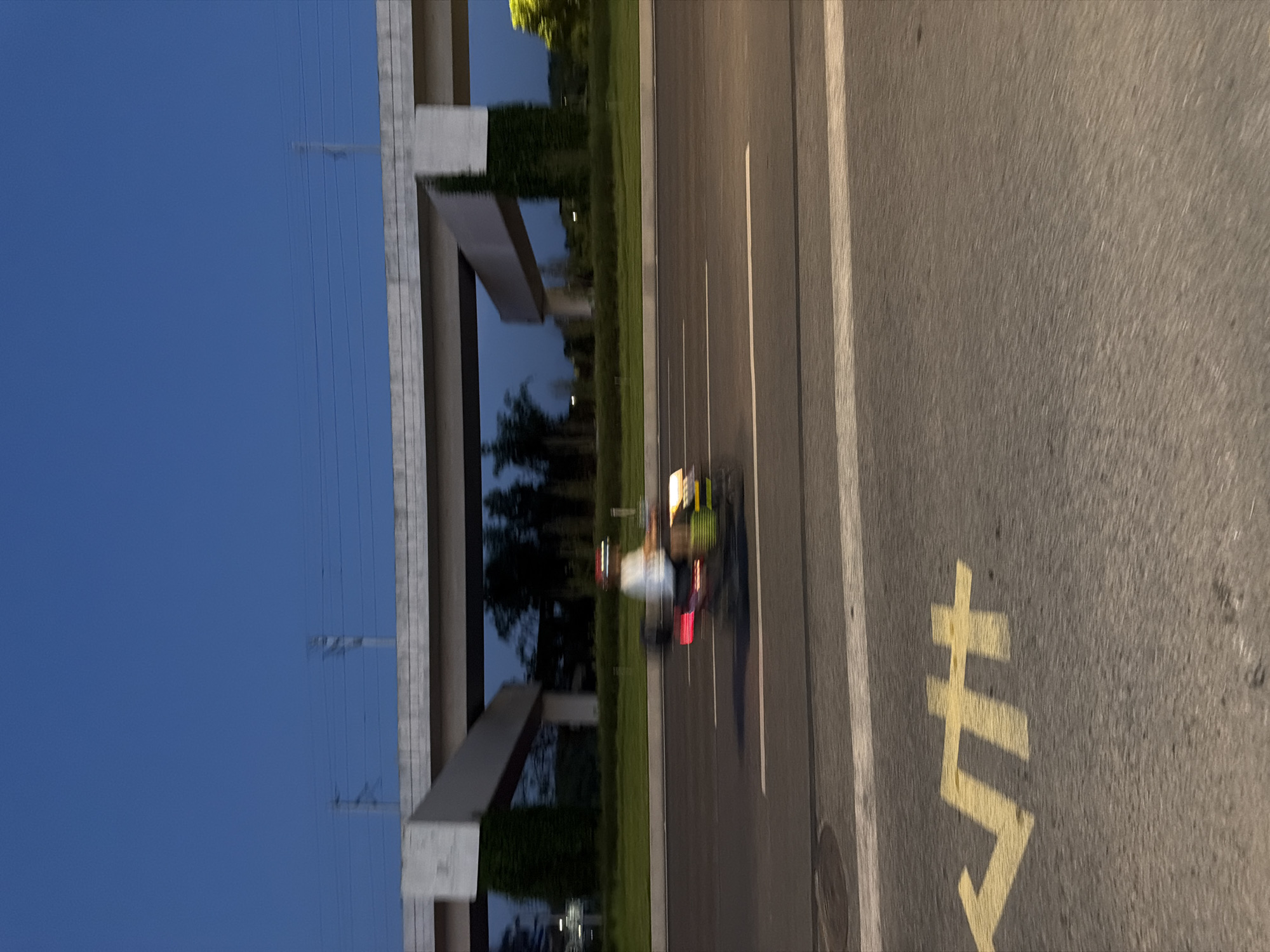 The image depicts a blur of motion on a road in Jiangjin District, Chongqing, China, likely at dusk due to the twilight sky. A person on a scooter, wearing a helmet and a light-colored shirt, is moving from left to right across the frame. The scooter has a bright yellow light on the front and red taillights. The road has white lane markings, and a yellow arrow marking is visible on the left side of the road. Overhead, a concrete overpass or bridge structure spans the scene, supported by pillars. Beyond the road, there is a strip of green grass and trees, with some darker foliage suggesting a park or natural area. Electrical wires and poles are visible above the overpass, against the deep blue sky. The overall impression is one of urban infrastructure and movement.