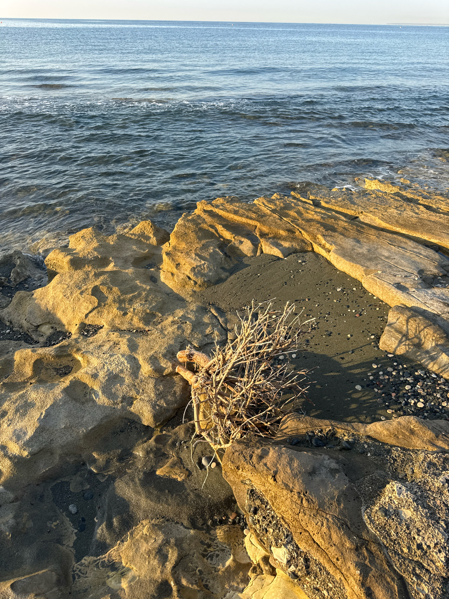 This is a vertical, eye-level shot of a rocky coastline in Agios Tychonas Community, Cyprus, at dusk or dawn, with the sun casting warm golden light on the scene. The foreground is dominated by textured, beige-colored rocks with indentations and shadowed hollows, interspersed with patches of dark, wet sand and small, smooth pebbles. A cluster of dry, light-brown twigs lies nestled in the sand within a rocky crevice. Beyond the rocks, the dark blue sea stretches to the horizon, with gentle waves lapping at the shore, creating a foamy white line where they meet the land. The sky above is a pale, hazy blue. There are no people or animals visible in the image.