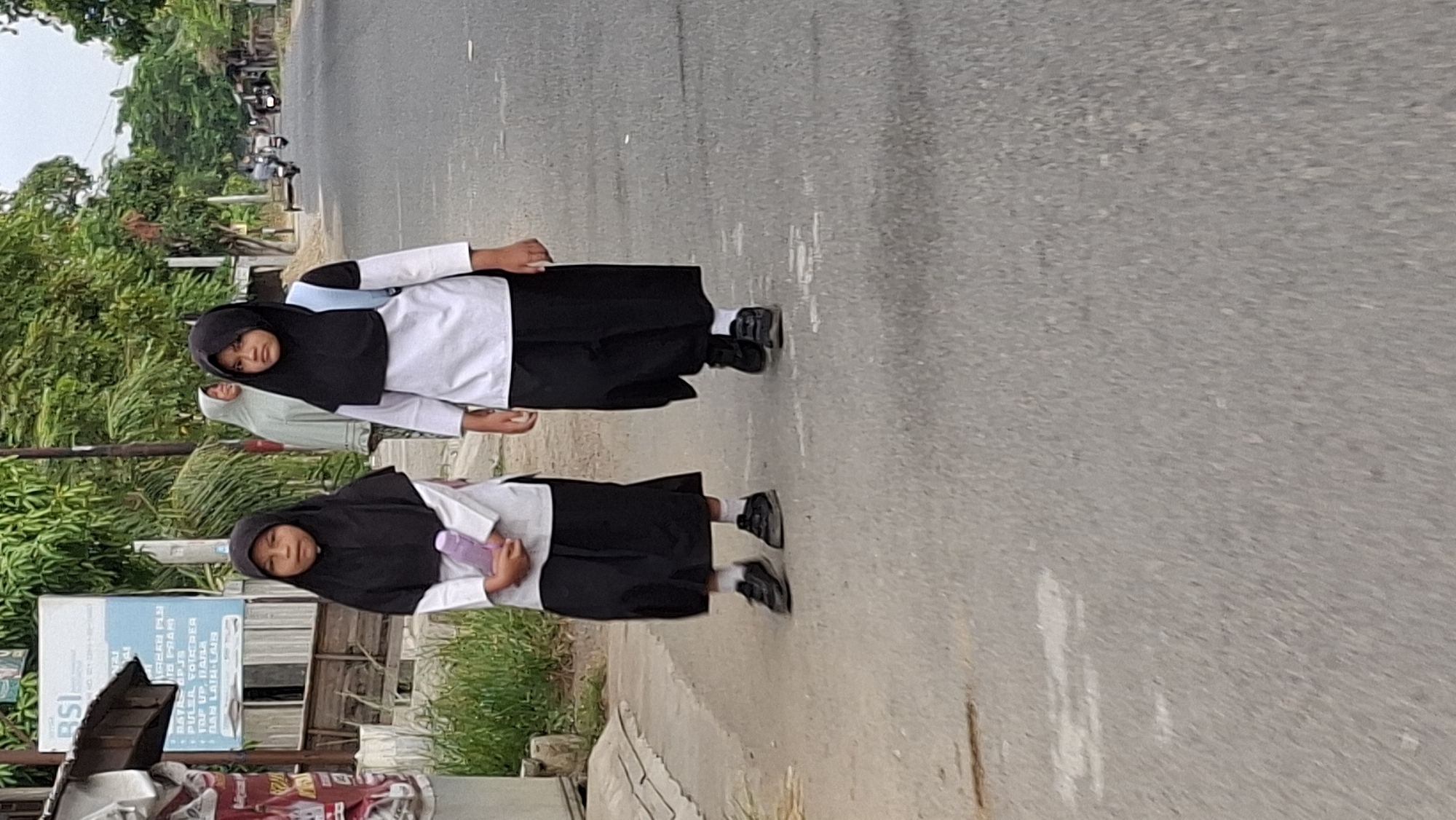 Two young girls, likely students given their matching uniforms, walk along a paved road. Both are wearing black hijabs and long black skirts over white long-sleeved shirts. The girl on the left is holding a light purple water bottle. The girl on the right has a light blue backpack on her shoulders. They are walking away from the viewer, towards the right side of the frame. The road is bordered by a grassy area on the left, with a building and some trees in the background. On the right side of the road, there are more trees and buildings, and a few motorcycles are visible further down the road. This scene appears to be in a suburban or semi-rural area of Lhokseumawe, Indonesia, possibly near a school.