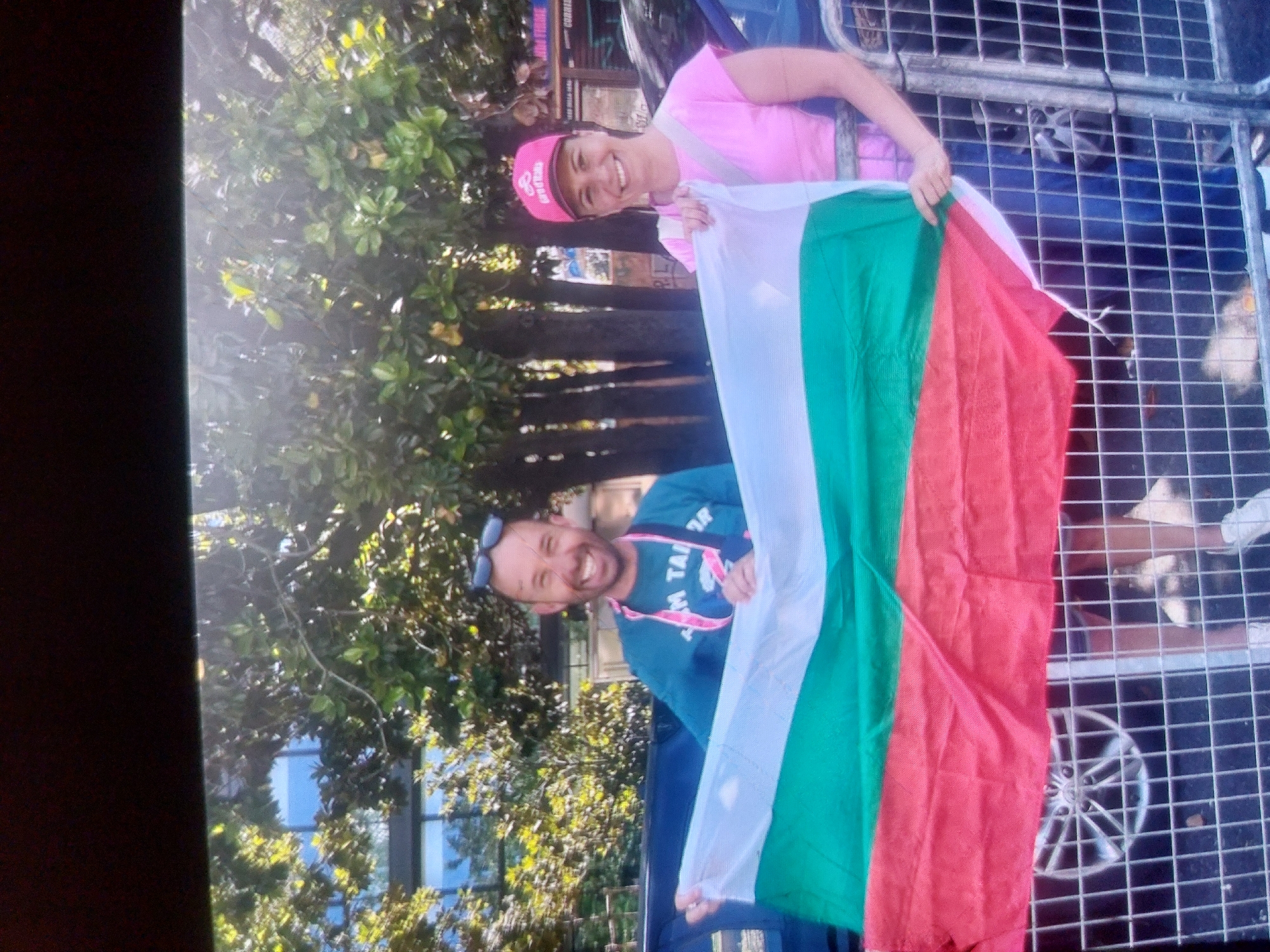 Two smiling individuals are holding the Bulgarian flag. The woman on the right is wearing a pink t-shirt and a pink baseball cap. The man on the left is wearing a teal t-shirt and sunglasses on his head. They are standing in front of a metal barricade. Behind them are trees and a building. The presence of the Bulgarian flag suggests a location within Bulgaria, potentially related to an event or celebration. More precise geolocation within the city of Tsarevo is not possible with the data.