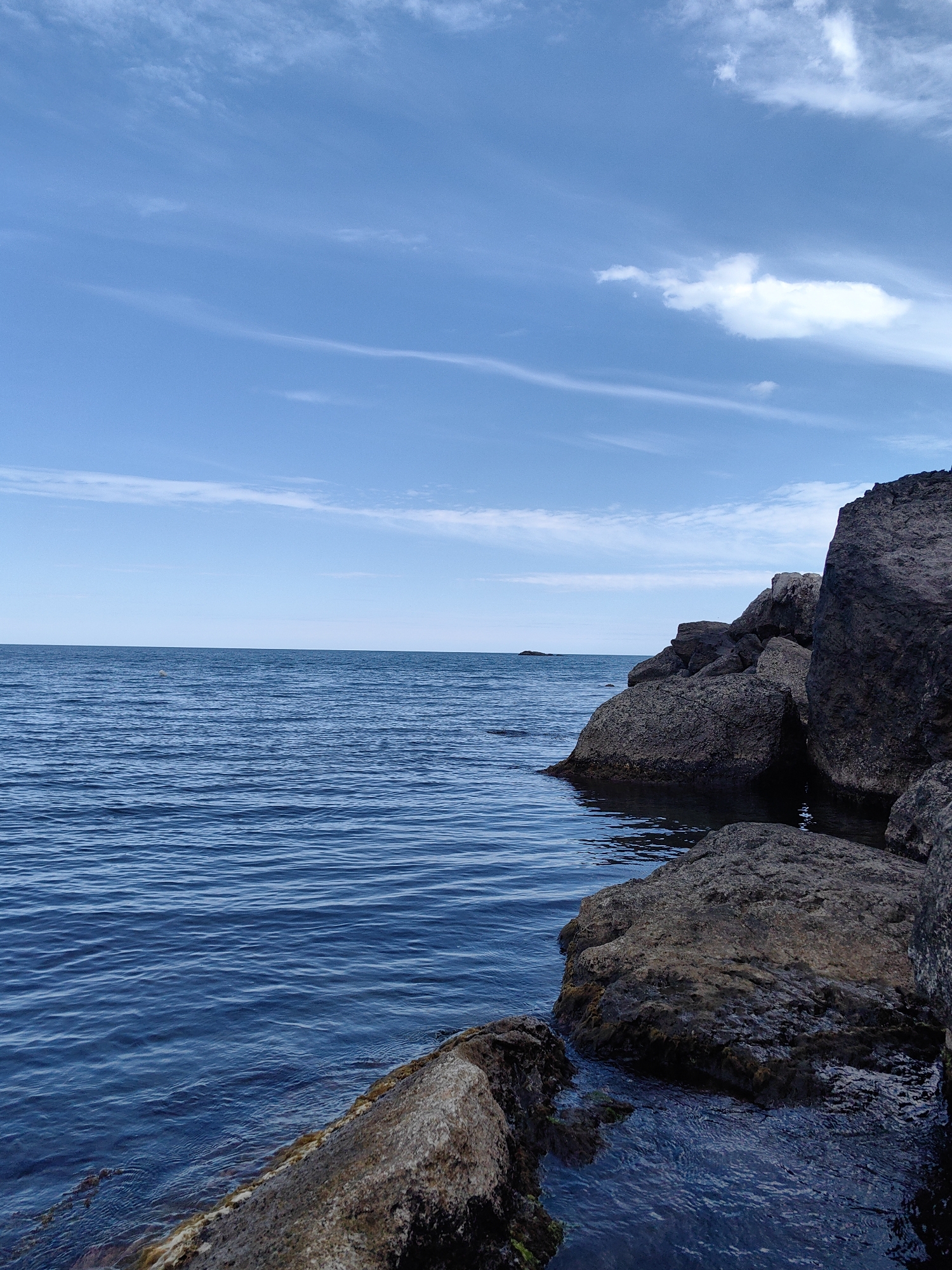 A vertical, medium-shot, outdoor, daytime photograph depicts a rocky coastline meeting the sea under a partly cloudy sky. The foreground features several large, dark, rough-textured rocks that extend into the deep blue water. The water is calm with gentle ripples, and its color deepens to a darker blue as it recedes toward the horizon. The horizon line is clearly defined, separating the sea from a pale blue sky that is streaked with thin, wispy white clouds. The overall impression is one of natural, rugged beauty. The location of Utios, Ukraine is not evident from the image itself.