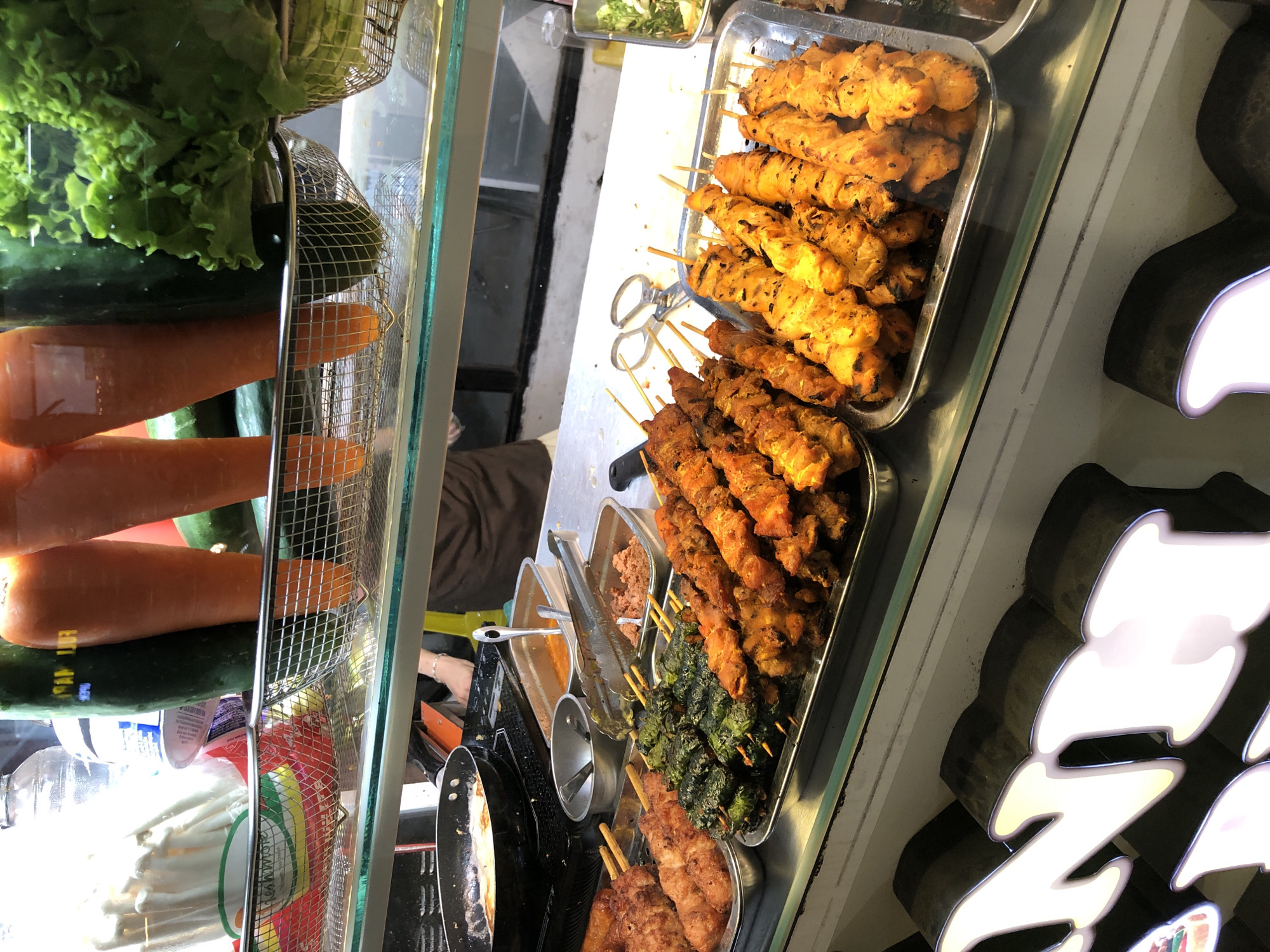 This image depicts a food stall in the city of Đà Nẵng, Vietnam. The stall displays a variety of grilled meat skewers, including what appear to be chicken and pork. These skewers are arranged on metal trays in the foreground. Behind the skewers, there are several stainless steel containers holding sauces or other food items, and a frying pan on a burner. In the background, large carrots and cucumbers are visible, along with a head of lettuce. A person, partially visible, is tending to the food. The overall environment suggests a busy street food vendor.