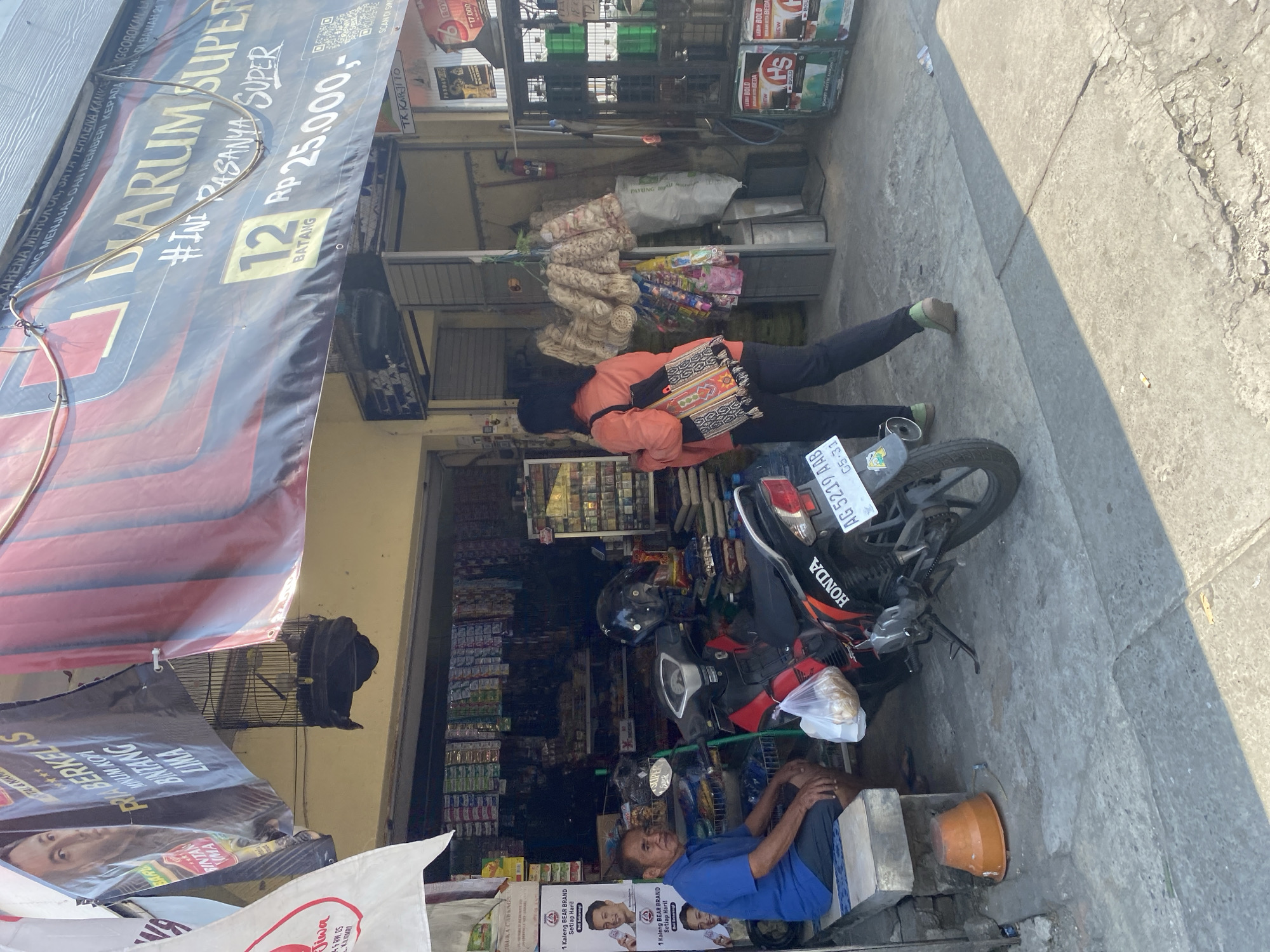 In Kediri City, Indonesia, a man sits on a low concrete bench outside a shop, wearing a blue short-sleeved shirt and dark shorts. A motorcycle is parked beside him, with a black helmet resting on its seat and a plastic bag containing food items hanging from its handlebars. In front of the bench, an orange bucket is placed on the ground. Behind the man, the shop displays various goods on shelves and in a glass cabinet, including cigarettes. A woman in an orange shirt and black pants walks away from the shop, carrying a bag over her shoulder. Several banners are displayed above the shop entrance, one of which features a large advertisement for "Djarum Super" cigarettes, including a QR code and pricing information. Another banner shows a close-up of a person's face and text in Indonesian. A birdcage with a bird inside hangs from a cable. The scene takes place on a sunlit street with concrete pavement and the building appears to be a small retail establishment.