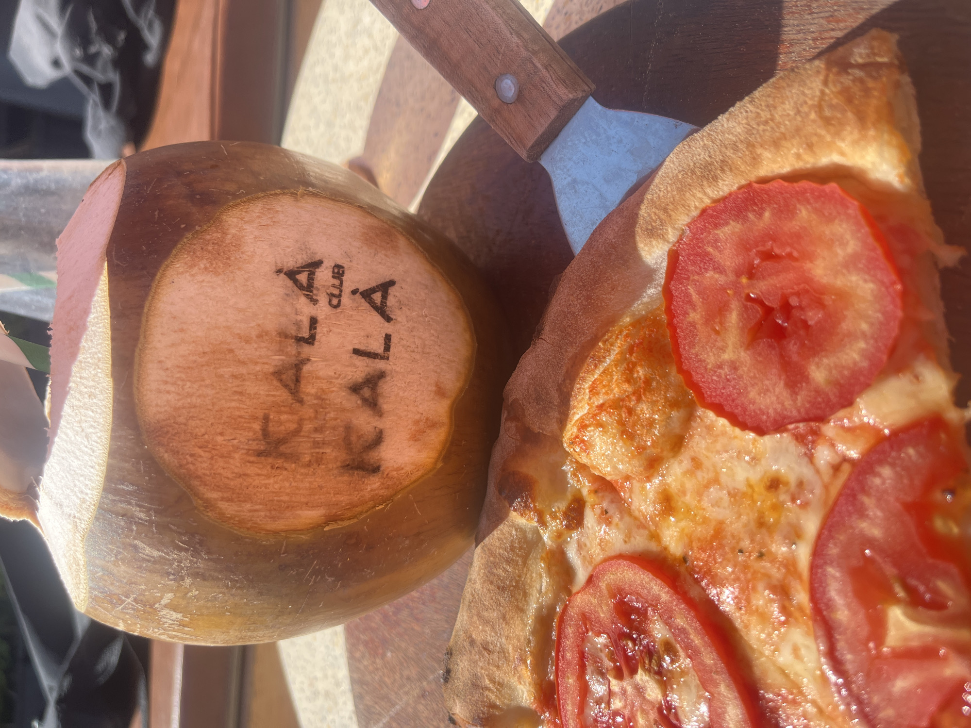 The image captures a close-up, top-down view of a single slice of pizza positioned next to a fresh young coconut. Both items rest on a rustic wooden surface, possibly a serving board.
The pizza slice features a golden-brown crust with a slightly charred edge, indicating a well-baked base. It is topped with melted white cheese and several round slices of vibrant red tomato. A pizza spatula with a wooden handle and a metallic blade is visible, tucked between the pizza and the coconut, suggesting the pizza has just been served or is about to be eaten.
The coconut has its top cut off, exposing its inner surface. Clearly branded onto this flat surface are the words "KALA CLUB" and "KALA" below it, indicating a specific venue or brand.
The scene is brightly illuminated by natural sunlight, casting strong highlights and distinct shadows, which suggests it is daytime, likely outdoors or in a well-lit open-air setting. The combination of pizza and a fresh coconut in such a sunny environment points to a leisurely dining experience, consistent with a holiday or resort setting in Đà Nẵng, Vietnam.
