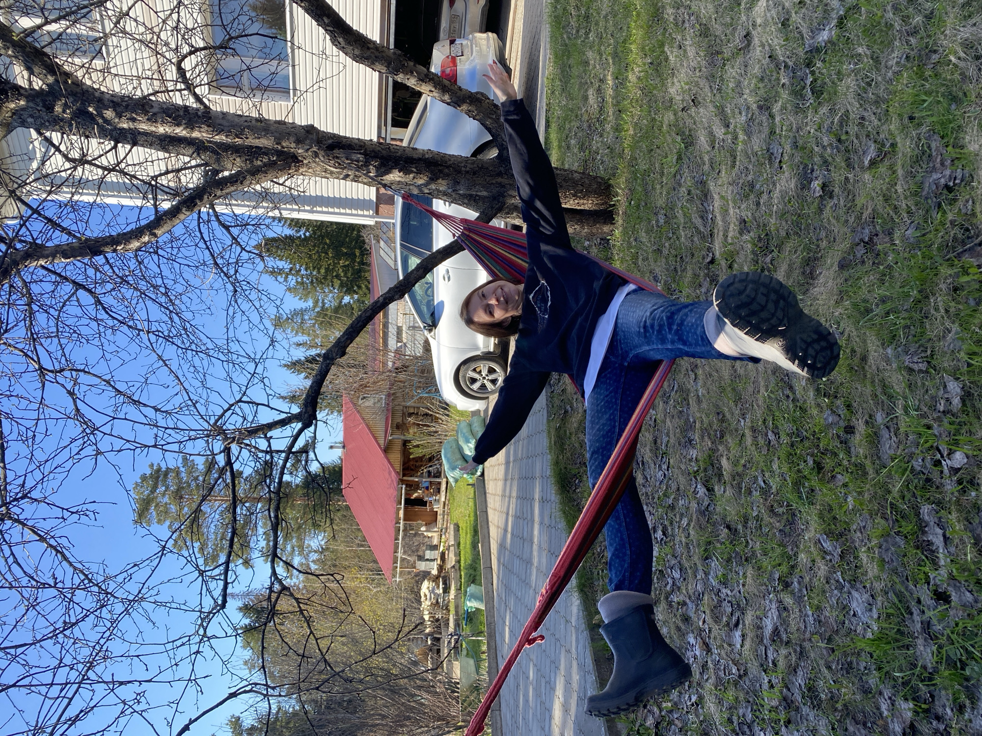 A woman is lying in a colorful hammock strung between two trees on a partly grassy area near a paved pathway. She is wearing jeans, a black hooded jacket, a white shirt, and black boots with gray socks. Her arms are outstretched. Behind the trees and hammock, there are houses and cars. A red-roofed building is visible to the left of the frame, partially obscured by tree branches. The sky is blue. The location is Прикамский, Russia.