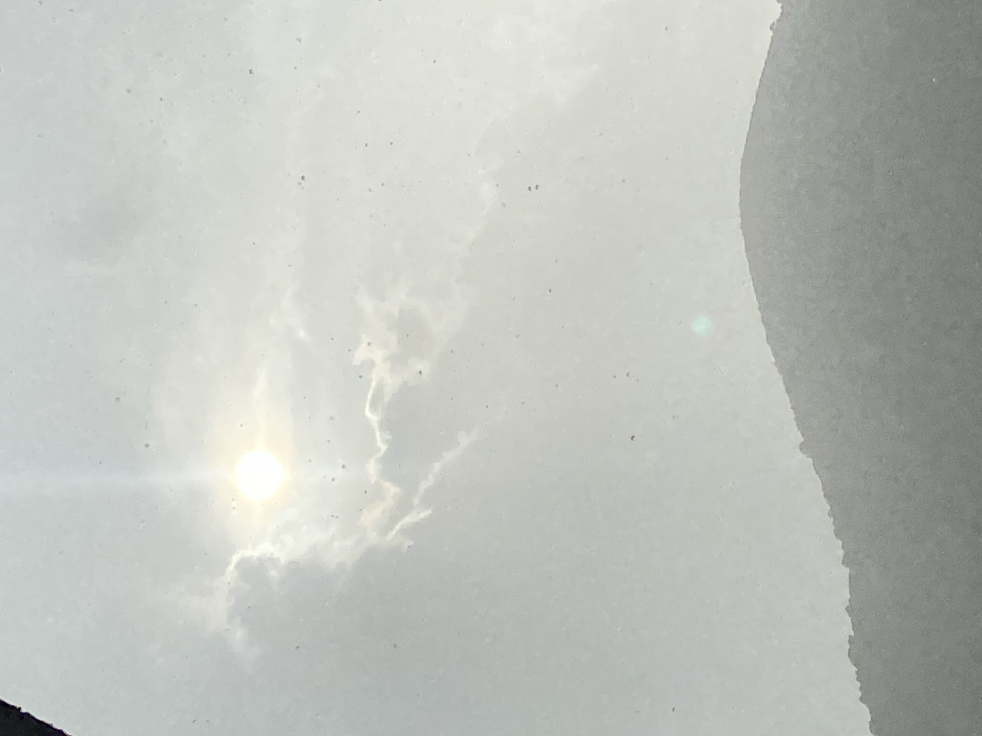 The image shows a bright sun partially obscured by clouds, with a hazy atmosphere. In the foreground, the silhouette of a rounded hill is visible against the sky. The overall scene suggests an outdoor environment, possibly a landscape view. There are no discernible people, objects, or specific actions occurring in the image. The location in Budva, Montenegro is not explicitly identifiable from the visual information present in the photograph.
