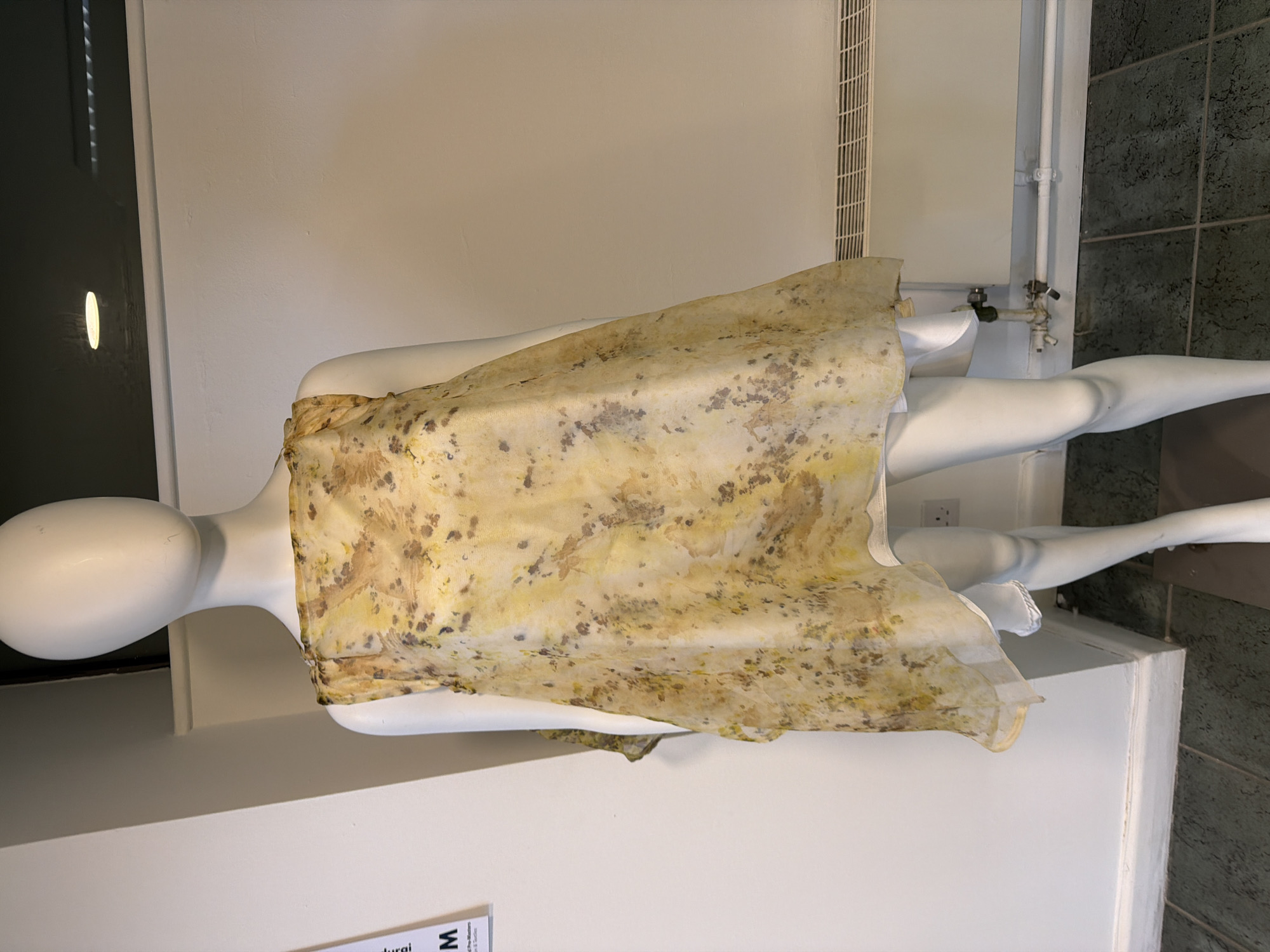A mannequin stands in what appears to be an indoor display, its body covered with a light, patterned fabric. The mannequin is positioned against a plain white wall, and there is a ventilation system and pipes in the background. The location is Epsom and Ewell, United Kingdom, indicated by the word "durqi" on a sign near the bottom of the photo.