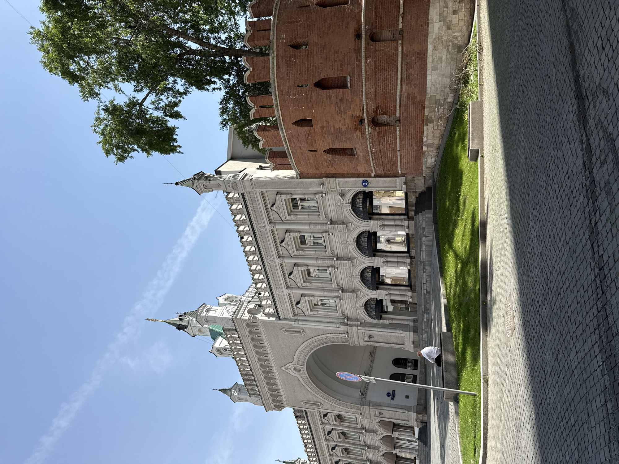The image shows a view of a street in Moscow, Russia. A historical building with white facade and intricate architectural details stands alongside a brick tower that is part of the Moscow Kremlin wall. A green tree is partially visible in the upper-left corner of the image. A patch of green grass separates the building from the cobblestone pavement on the right. A person is bent down on the side of the road. The sky is clear and blue.