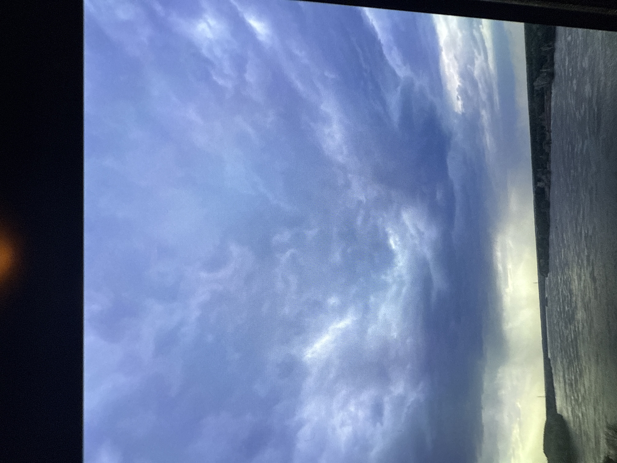The image shows a landscape oriented view of a cloudy sky and water, presumably a river or a large body of water. The clouds are varied in density and coloration, ranging from a light grey/white to a darker blue-grey. The water appears calm, with its color also varying from light to dark grey. The photograph's location is unknown and no people, interactions or specific actions can be identified.