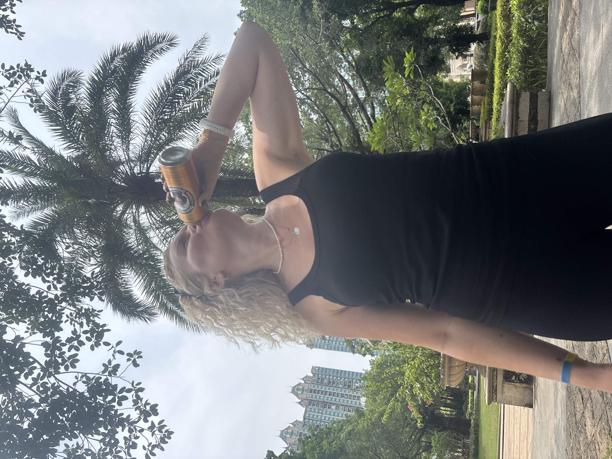 A woman with blonde curly hair is drinking from a beer can outdoors. She is wearing a black tank top, black leggings, a pearl necklace, and a white watch. A blue and yellow bracelet is visible on her left wrist. The background shows palm trees, lush green foliage, and modern high-rise buildings. The scene appears to be in a park located in the Nanhai District of China.