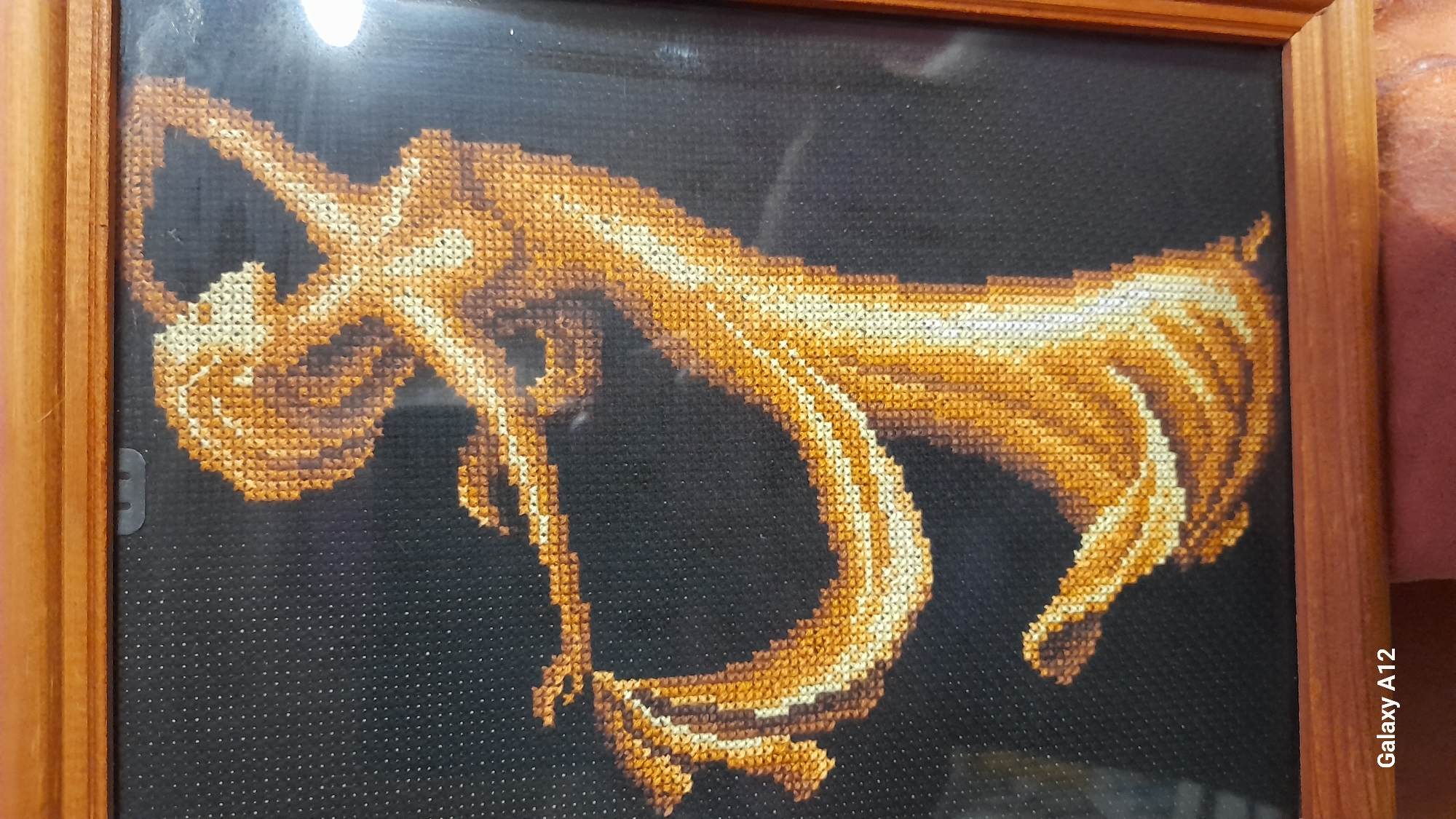 A framed cross-stitch artwork depicting a stylized golden figure on a black background. The figure appears to be dancing or in motion, with flowing lines and curves. The frame is made of wood. The location is not visible from the image.