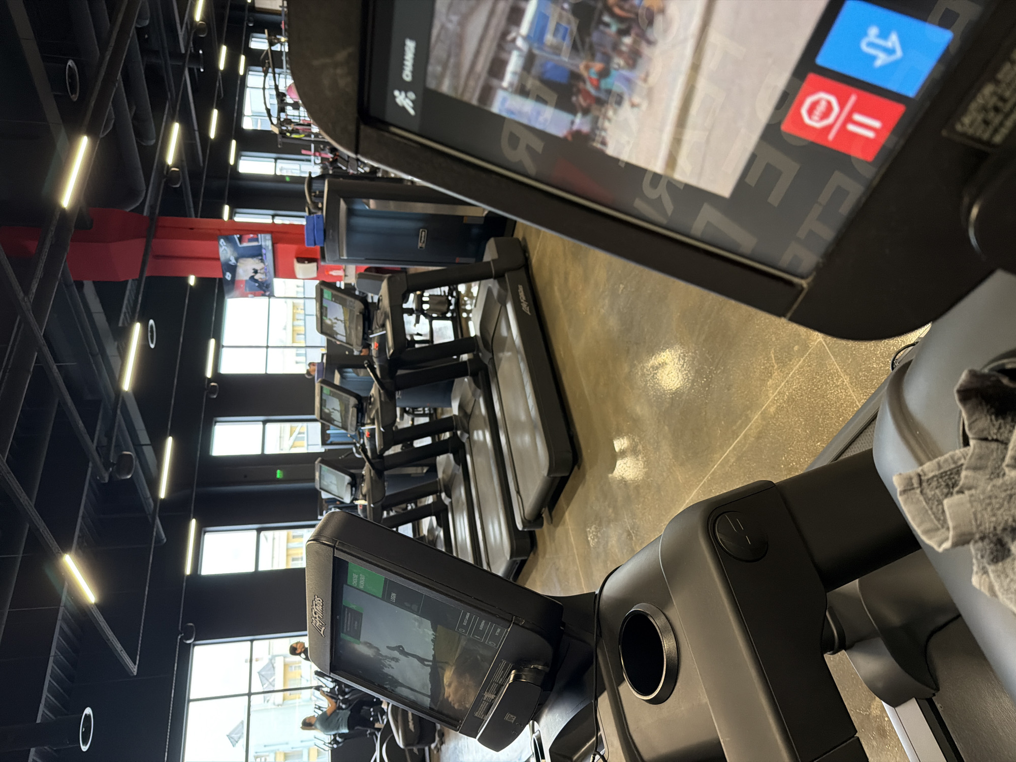 The image shows the interior of a gym, possibly located in Plovdiv, Bulgaria. A row of Life Fitness treadmills are positioned in the foreground and middle ground, facing towards windows that reveal an urban landscape. The gym features a high ceiling with visible black metal framework and recessed lighting. A red structural column with a screen displaying video content is visible in the background. The floor is a neutral gray tone. The overall scene suggests a modern fitness facility.