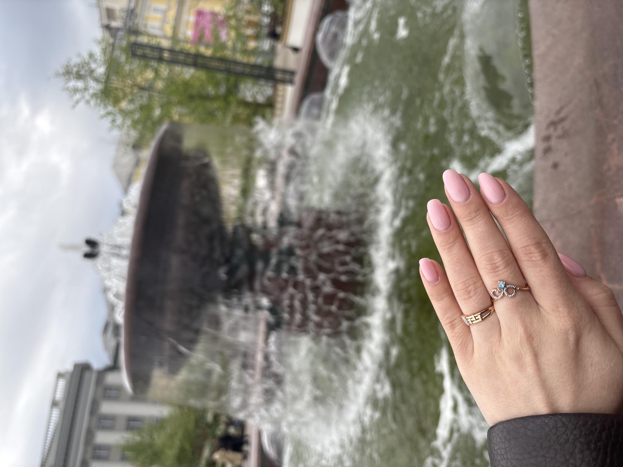 A woman's hand wearing two gold rings is in the foreground with a fountain in the background. The hand is fair-skinned and has manicured pink nails. One ring is a simple band and the other has a small blue stone. The fountain is large and multi-tiered, with water cascading down. The background is blurred but appears to be a city scene with buildings and trees. The location is identifiable as Moscow, Russia, due to the architecture and style of the fountain.