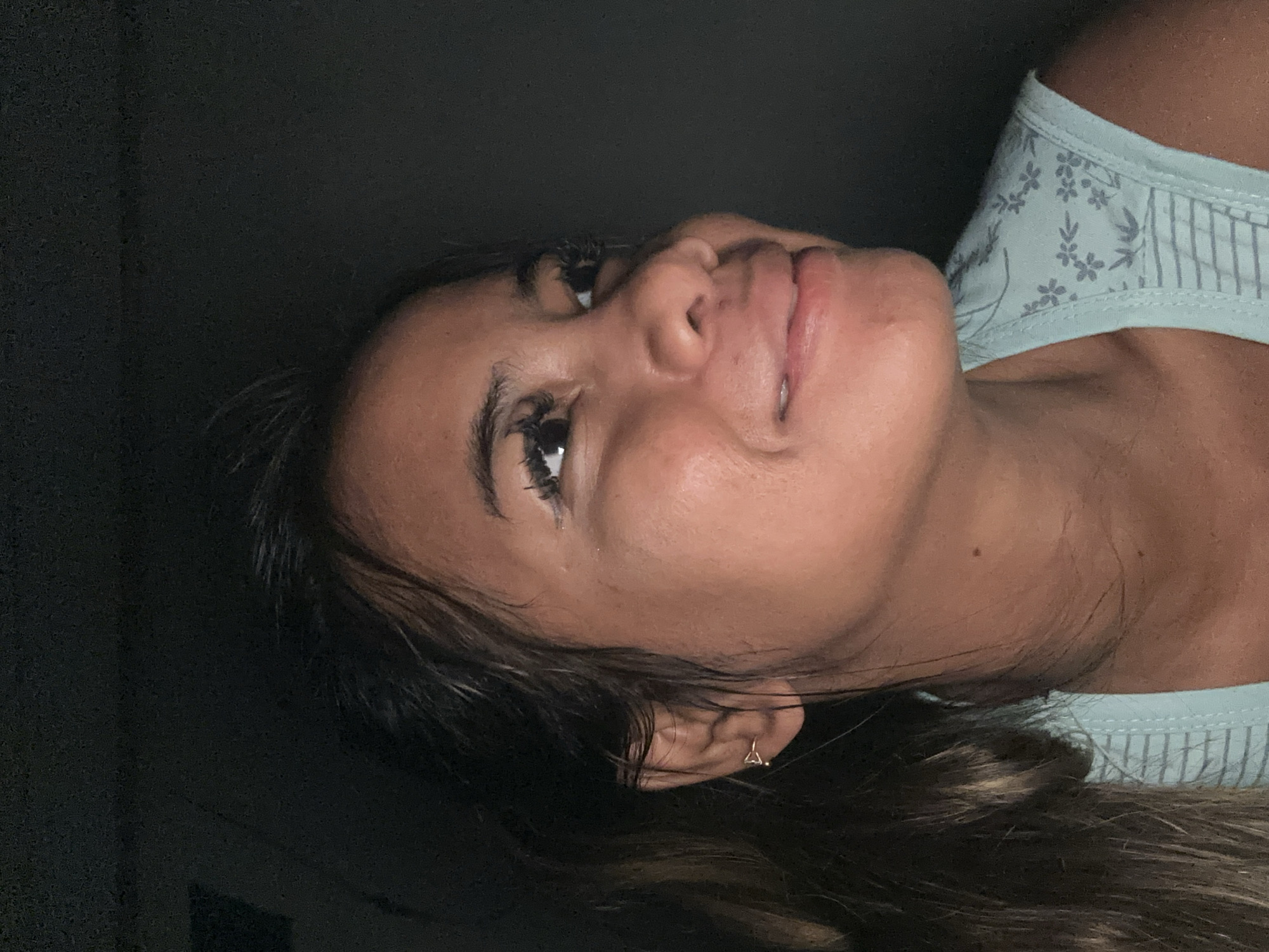 A young woman is captured in a close-up, horizontally oriented shot, appearing to be resting with her head tilted to the side. Her dark hair frames her face, with some strands falling over her shoulder. She exhibits a subtle smile or smirk, with her lips slightly parted, and her eyes are open, looking upwards towards the upper right of the frame. Her complexion is warm, and her eyelashes appear long and dark.
She is wearing a light teal or mint green top, possibly a tank top or camisole, featuring a delicate pattern of light grey floral elements and stripes. A small, golden, heart-shaped stud earring is visible in her right earlobe. The lighting is dim, casting soft illumination on the left side of her face, while the background remains uniformly dark and out of focus, suggesting an indoor setting. The overall mood is relaxed and intimate, likely captured during the evening or night in North Jakarta, Indonesia. No discernible text is visible in the image.