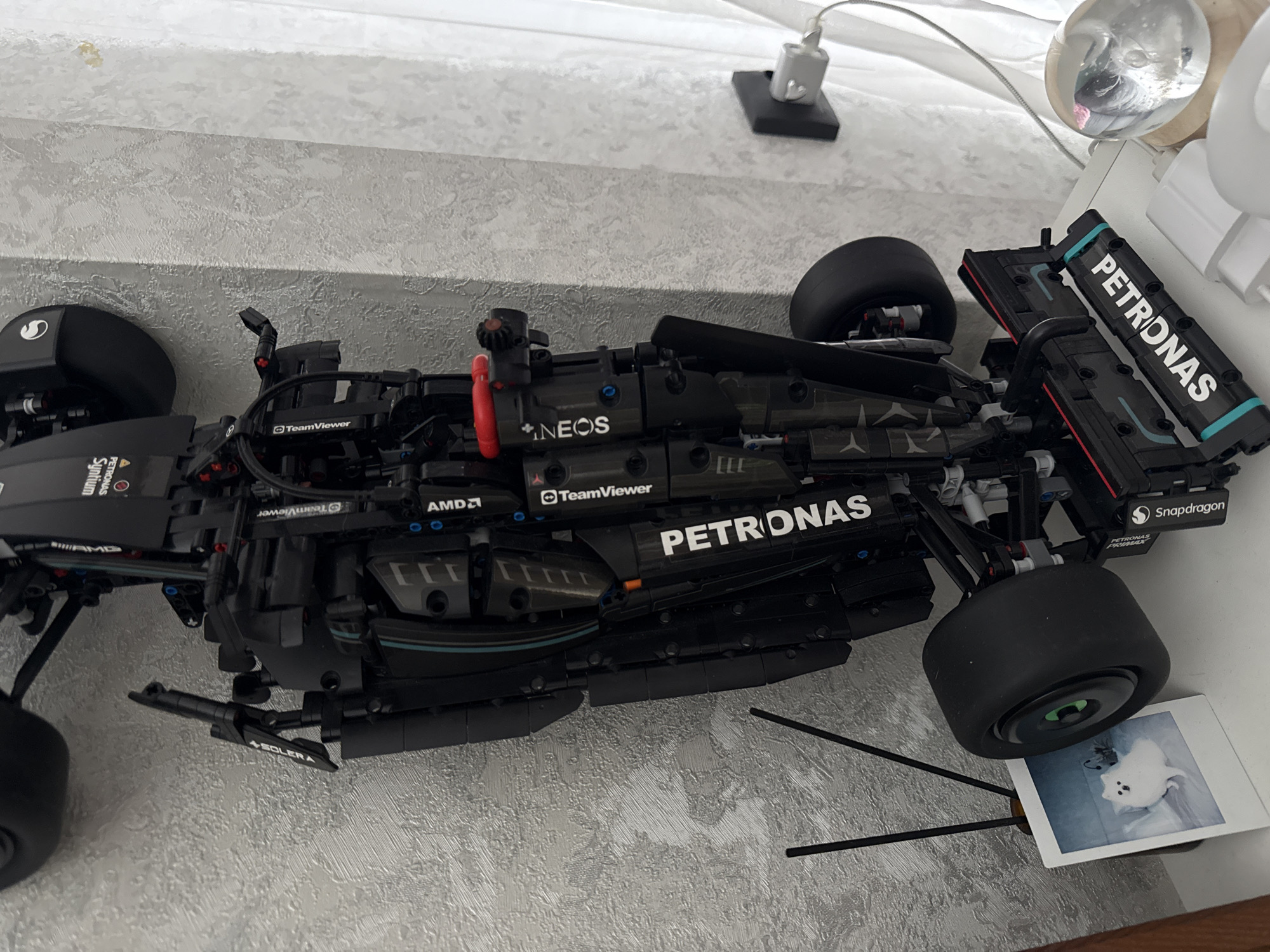 The image displays a detailed black Lego Technic model of a Mercedes-AMG F1 W14 E Performance race car, positioned on a light-colored, textured surface, likely a desk or shelf, against a patterned wall, suggesting an indoor setting in Лобаново, Russia. The car model is angled, showcasing its intricate mechanical structure and various sponsor logos prominently.
Visible text on the car includes "PETRONAS" (on the side, rear wing, and other areas), "TeamViewer" (multiple instances), "NEOS" (referring to INEOS), "AMD" (repeatedly), and "Snapdragon". Partially visible text includes "Syntium" and "SOLERA". The car's tires are black and smooth, characteristic of racing slicks.
In the background, a bright, diffused light source, possibly a window, illuminates the scene from above. Electronic accessories such as a silver charging adapter and the base of a lamp are visible. A clear spherical object, likely a paperweight or decorative item, reflects a distorted image of what appears to be a dog's face. Directly beneath the car's rear wheel, a small instant photograph is placed, clearly depicting a fluffy white dog. The overall impression is one of a meticulously built model displayed as a collector's item or personal decoration, incorporating subtle personal elements with the dog photos.