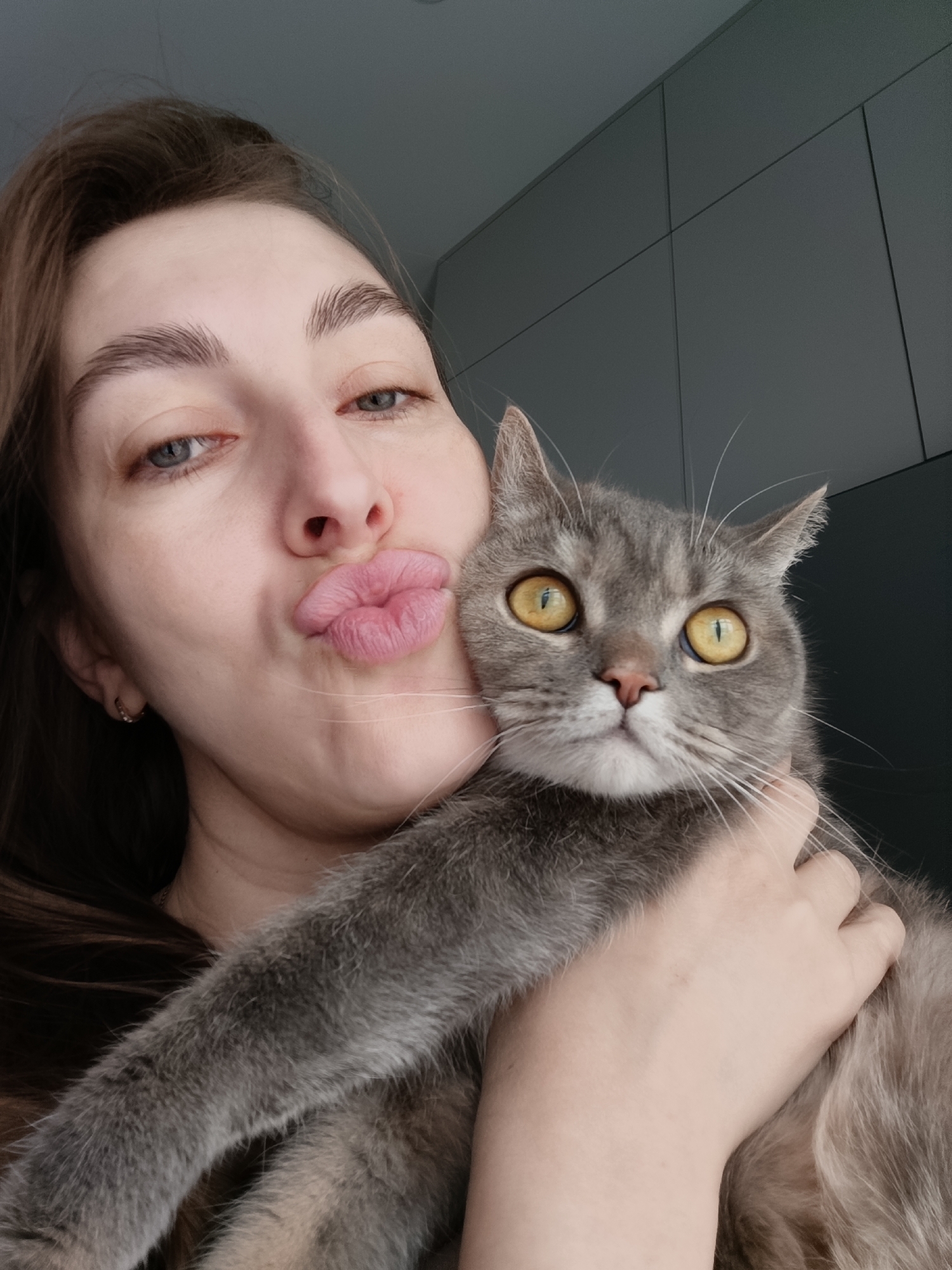 A woman is holding a grey cat. She is pouting her lips for a selfie. The background shows a dark grey wall, likely indoors. The location is Chaikovsky, Russia.