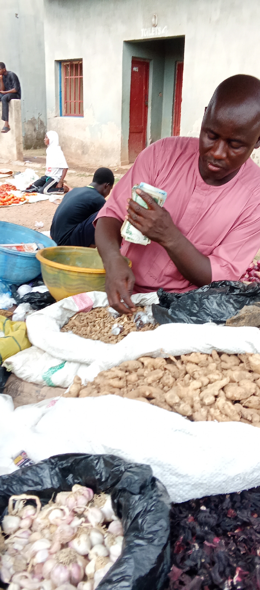 The image shows a vendor in a market in Akwanga, Nigeria, counting money. He is wearing a pink shirt and is surrounded by bags of ginger, garlic, and other dried herbs. There are other people visible in the background, and there is a building with the word "TOILETS" visible above a door.