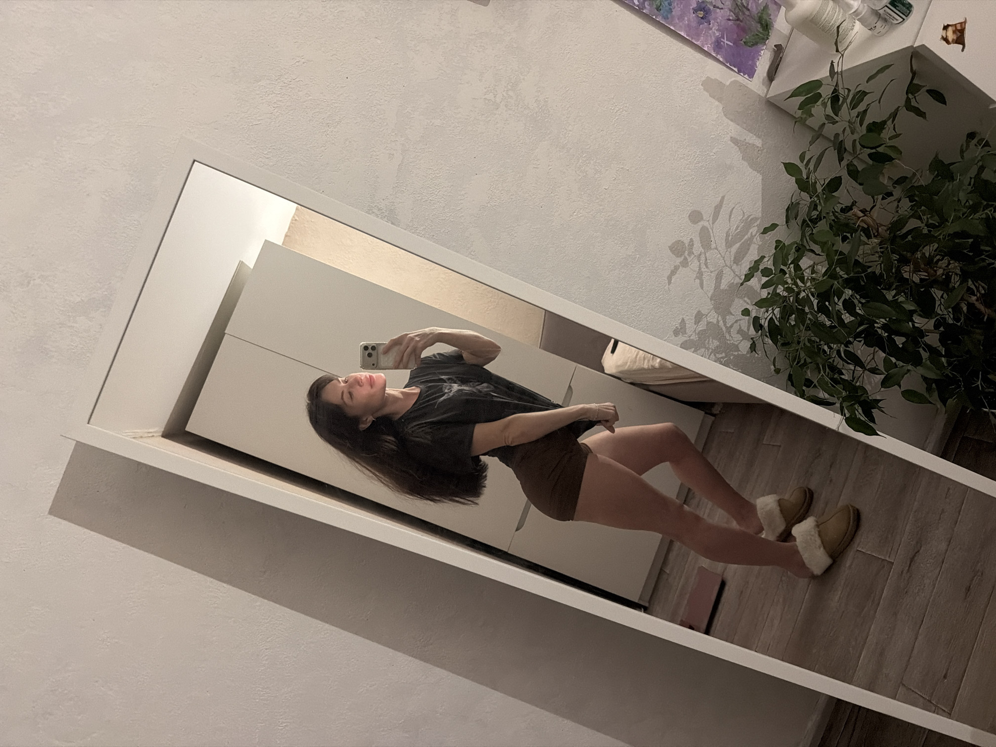 A woman stands in front of a mirror, taking a photo of herself with a smartphone. She is wearing a dark t-shirt and brown shorts, and slippers. The background includes a white closet, a houseplant, and a wooden floor. The image was taken in Perm, Russia.
