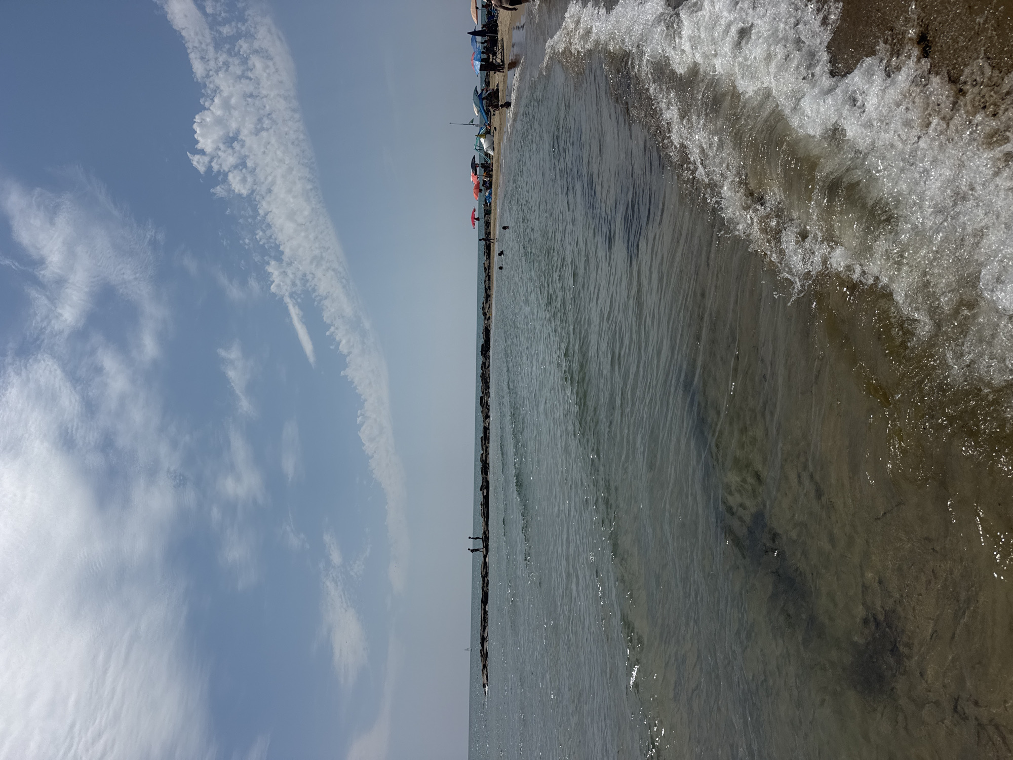 This image captures a sunny day at the beach in San Felice Circeo, Italy. The foreground shows the gentle waves of the sea lapping onto a sandy shore. In the midground, the calm water extends towards a man-made breakwater constructed of dark, irregular rocks. Two figures are visible standing on this breakwater, silhouetted against the horizon. Further down the coastline, a beach scene unfolds with several colorful umbrellas providing shade for beachgoers. People are scattered across the sand and in the shallow water. The sky above is a clear blue, punctuated by wispy white clouds. The overall scene depicts a relaxed atmosphere typical of a summer day at the seaside.